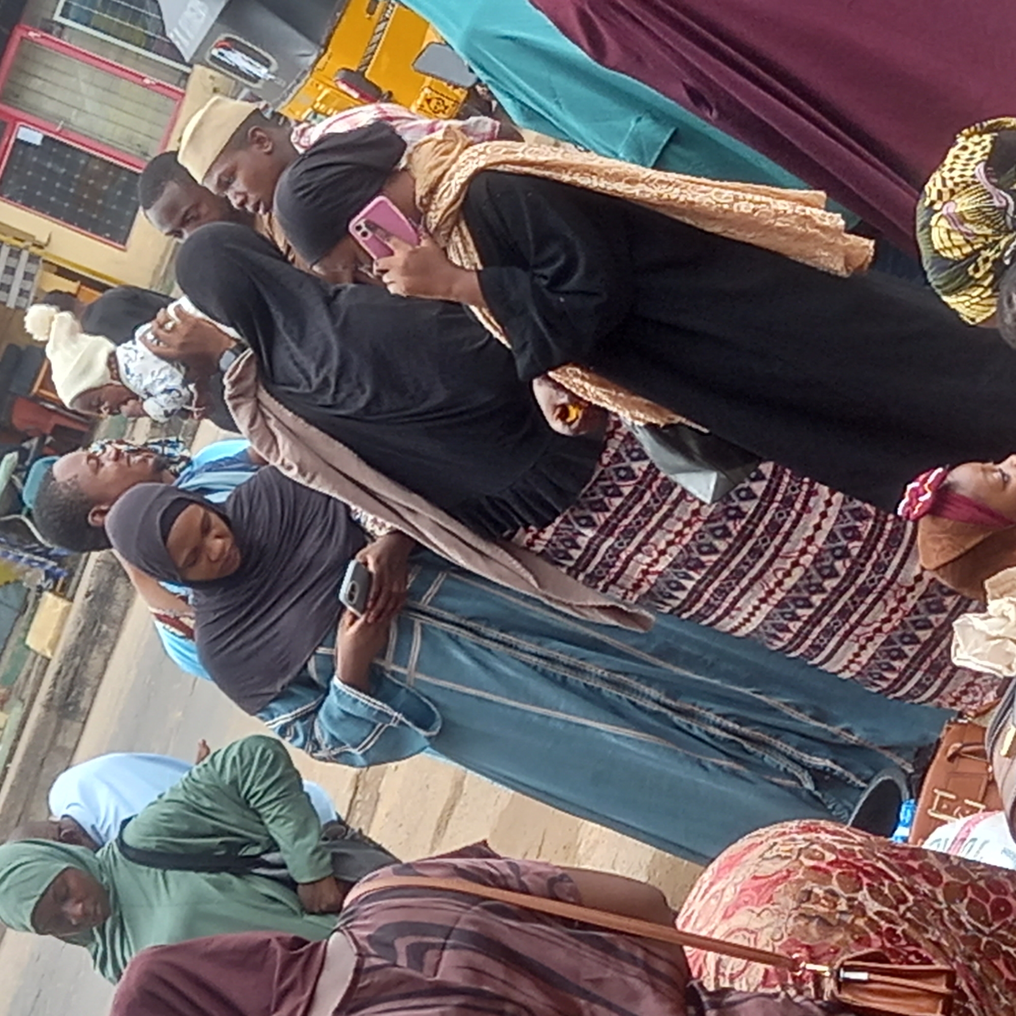The image appears to be taken in Sardauna, Nigeria, showing a group of people standing or sitting close together. Many of the individuals are wearing traditional Muslim clothing, including hijabs and robes of various colors such as black, blue, green, and maroon. Several people are looking at their phones. A man with a tan-colored hat is among the group. There is a yellow vehicle in the background, suggesting an urban environment. The setting appears to be on or near a street.