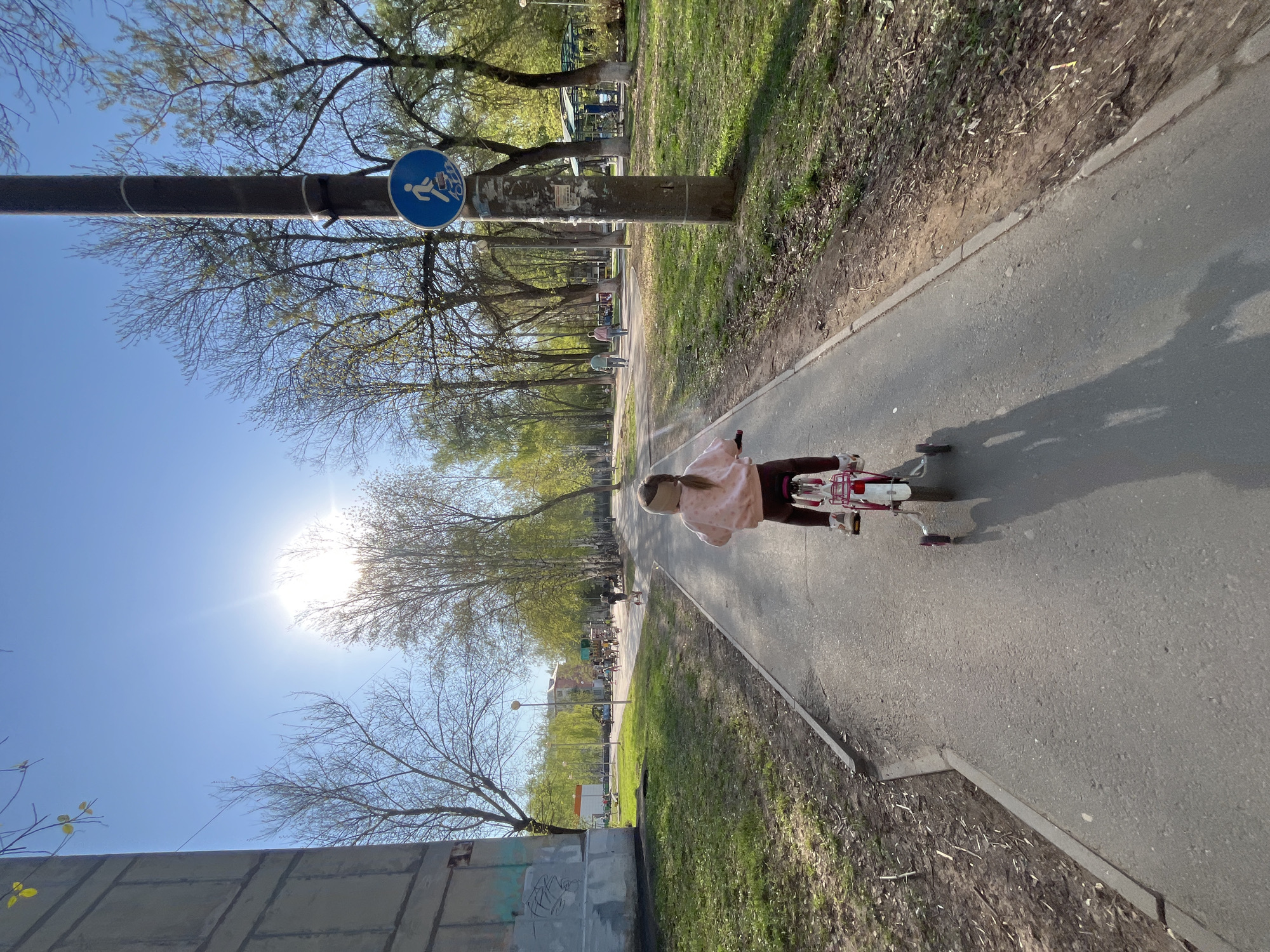 A child is riding a red tricycle on a paved pathway in what appears to be a park. The pathway is flanked by grassy areas and trees. A sign indicating a cycle path is visible attached to a pole. In the background, other individuals can be seen walking along the path. The environment is bathed in bright sunlight. The image was taken in the city of Izhevsk, Russia.