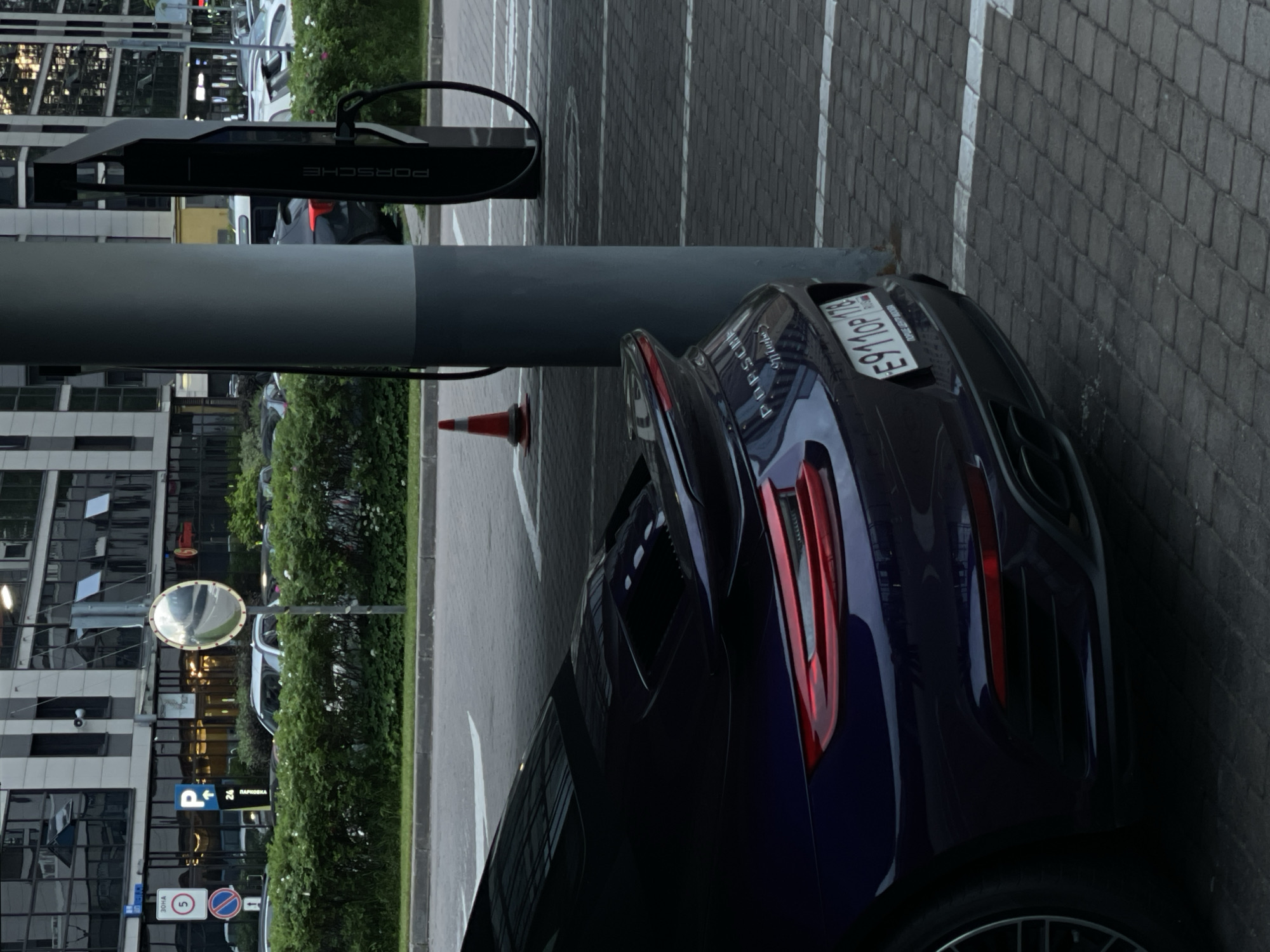 In an outdoor urban setting, a dark blue Porsche sports car is parked in front of a Porsche electric vehicle charging station. The car, visible from its rear quarter, has a prominent spoiler and taillight design. The charging station, a tall black structure with "PORSCHE" branding, is positioned behind the car. A large, grey cylindrical pillar is situated between the car and the charging station. In the background, a modern building with large windows and a landscaped hedge line can be seen. A traffic cone is placed near the pillar. Parking lot markings are visible on the paved ground, which is made of interlocking bricks. The scene suggests a charging stop for an electric Porsche.