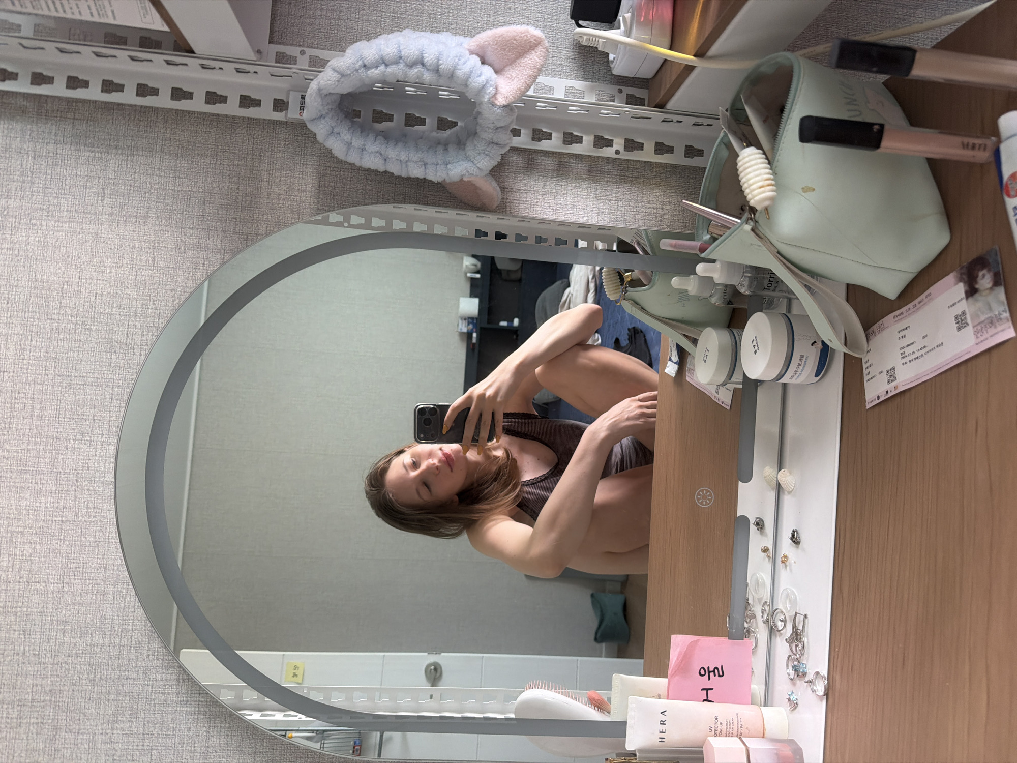 A young woman is taking a mirror selfie in a room in Dongducheon-si, South Korea. The woman is sitting on the floor with her legs crossed, holding a smartphone in her right hand. She is wearing a dark-colored tank top. Her hair is brown and falls past her shoulders. The room appears to be a bedroom or dressing area. In the foreground, a wooden vanity table is visible, cluttered with various beauty products. These include several tubes and jars of skincare and makeup, makeup brushes, and a light blue makeup bag with a white ribbed object sticking out of it. A light blue, fluffy headband with bunny ears is hanging on a white metal shelving unit to the right of the mirror. The mirror itself is oval-shaped and framed with a subtle decorative edge. In the reflection, the woman's face is visible, along with a portion of the room behind her, which includes a bed and shelves. The wall behind the woman and the shelving unit has a textured gray wallpaper.