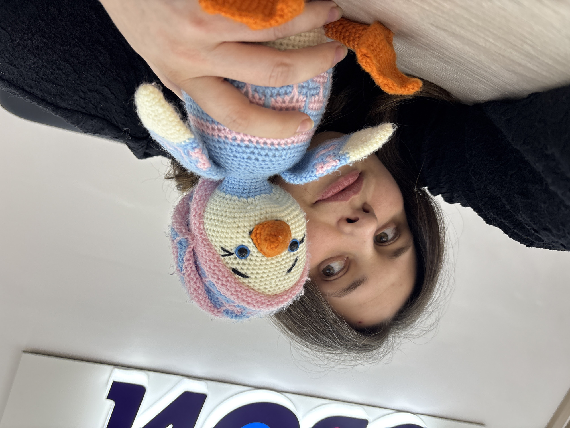 A woman with fair skin and shoulder-length dark hair with grey roots, wearing a black, textured garment, is holding a hand-made, crocheted toy penguin. The penguin has a cream-colored face, an orange beak, blue eyes, and a pink and blue striped hat. The penguin’s body is light blue with pink and white stripes, and it has cream-colored wings and orange feet. The woman's face is close to the penguin, and she is looking at the camera. A partially visible logo with purple and white shapes is in the lower part of the frame. The image was taken in the city of Chaikovsky, Russia.