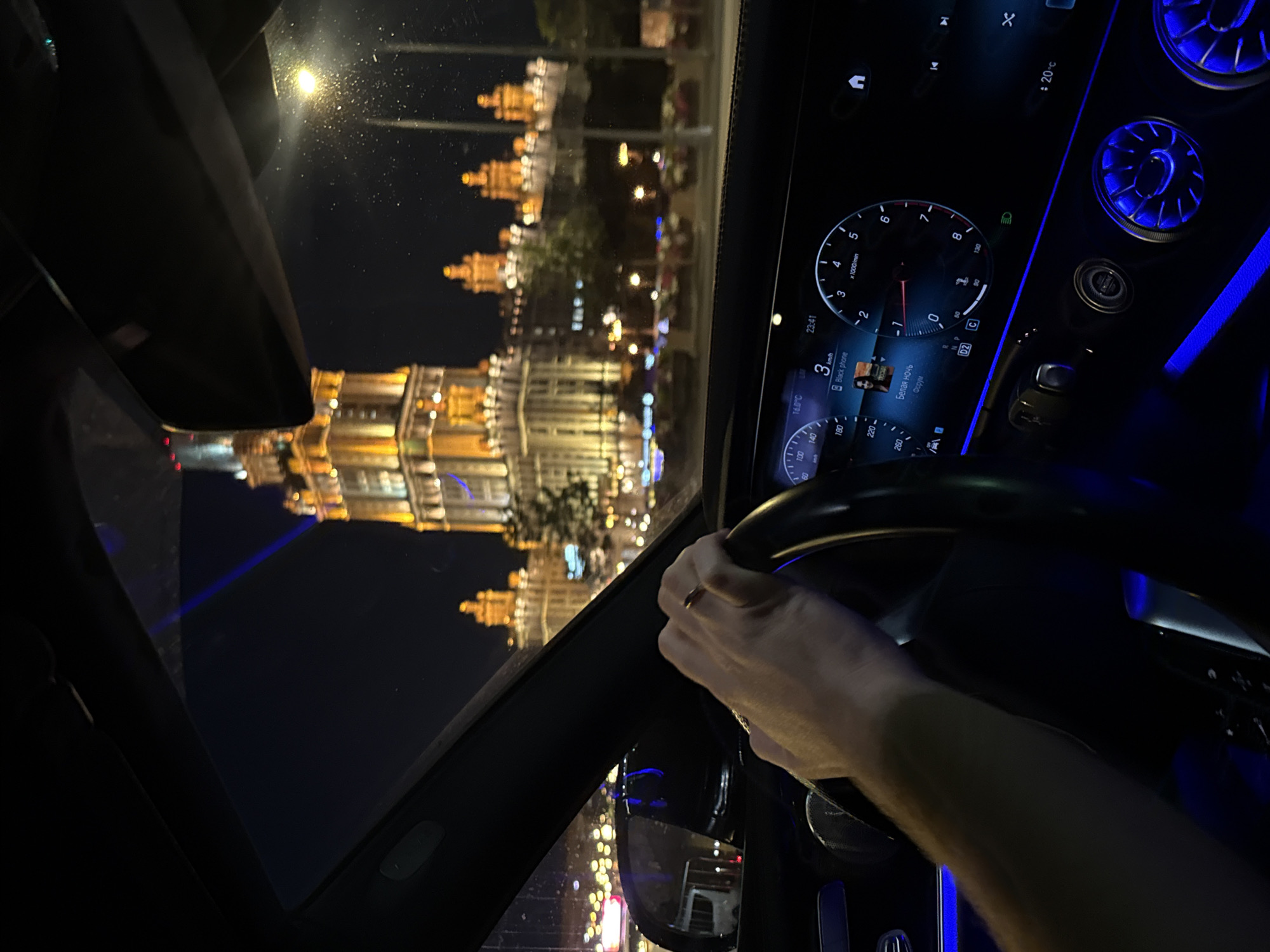 The image is taken from inside a car at night, looking out through the windshield. A person's left hand, wearing a ring, is on the steering wheel. The car's dashboard is visible, displaying information on a digital screen. Outside, a large, illuminated building with a prominent tower is visible, characteristic of Stalinist architecture. This building is likely Moscow State University, a landmark in the city. The surrounding area is lit by streetlights and the building's illumination, with other smaller illuminated buildings and trees visible in the background. The overall scene suggests a night drive through Moscow, Russia, with a view of a significant architectural monument.