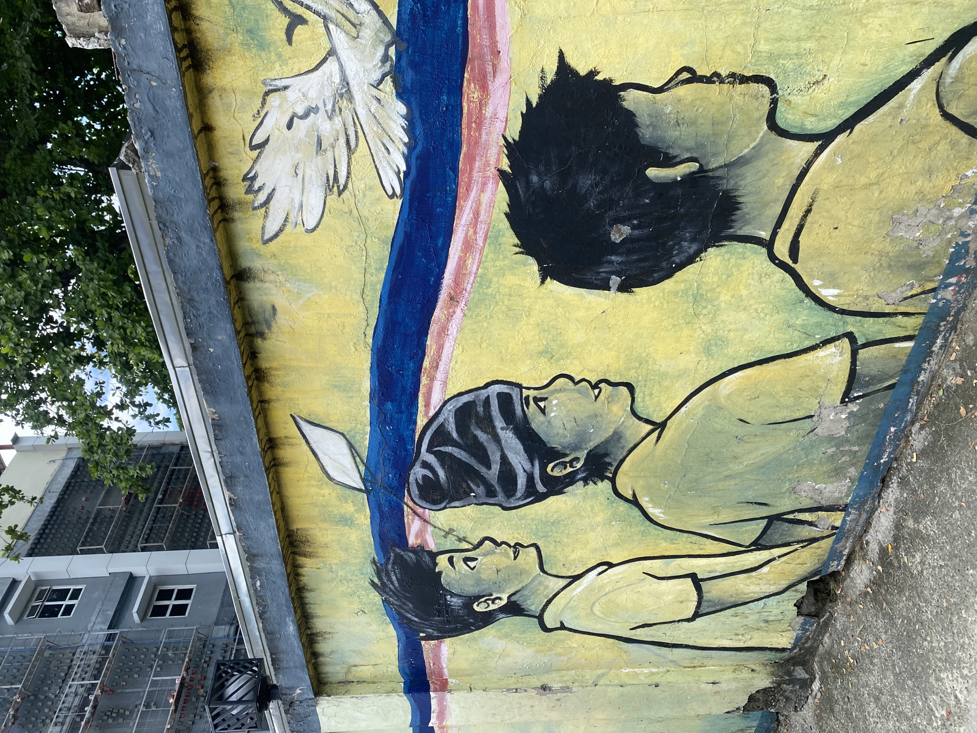 This media file presents an outdoor mural painted on a weathered concrete wall, captured from a slightly rotated, upwards-looking perspective. The mural depicts three stylized human figures, appearing to be youth or children, positioned vertically one above the other, all looking upwards towards the top of the frame. The figures are rendered with simple black outlines and filled with muted yellow and green tones for their skin and clothing. Two figures have dark hair, while the middle figure wears a striped head covering.
To the left of the figures, a white dove with large, outstretched wings is depicted flying upwards. Running vertically through the mural, between the dove and the figures, are two distinct stripes: one deep blue and the other a reddish-pink. The yellow background of the mural shows signs of age, with visible cracks and areas where paint has flaked off.
Beyond the mural wall, the scene includes elements of an urban environment. Lush green tree foliage is visible against a bright sky, indicating daytime. Below the trees, a modern multi-story building with a grey facade and grid-patterned elements is partially seen. The base of the mural wall meets a rough, paved ground surface. The upward gaze of the figures and the presence of the dove suggest themes of hope, aspiration, or peace. No discernible text is visible within the image. The setting appears to be a public outdoor space in a tropical or subtropical region, consistent with Davao City, Philippines.