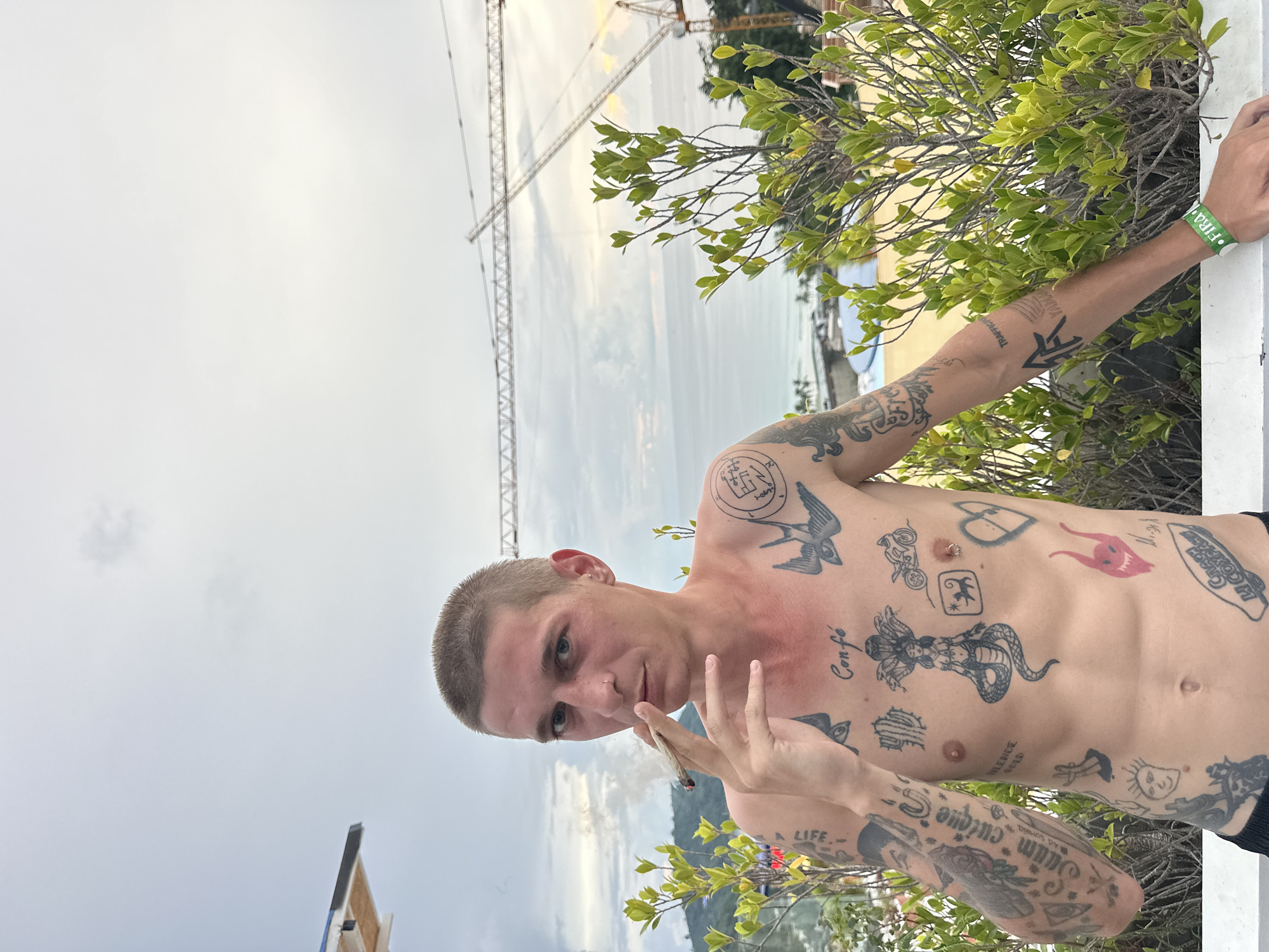A young, shirtless Caucasian male with short hair and a nose ring is standing outside, smoking a joint. He has numerous black tattoos on his chest and arms, including a snake woman, swallows, a motorcycle, a heart, and various script. He is looking directly at the camera. The background shows a cloudy sky, the ocean, and two construction cranes. Green foliage is visible to the right and left of the subject. The location appears to be Karon, Thailand.