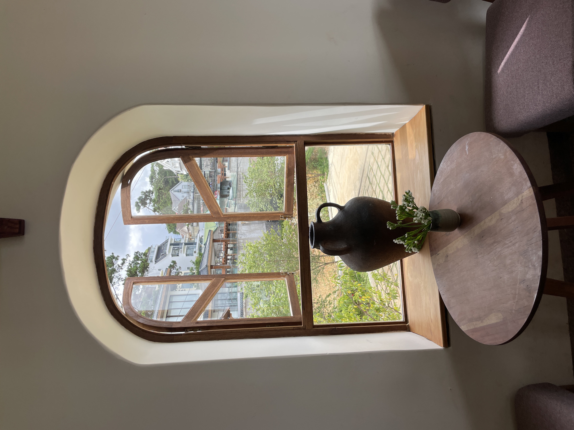 The image captures an interior scene with a large, arched window overlooking a city street in Da Lat, Vietnam. The window is framed by warm wooden trim and is partially open, revealing a view of multi-story buildings, trees, and a cloudy sky. On the windowsill, a dark, wide-mouthed vase stands prominently. In front of the window, a round, wooden table is set with a small ceramic vase containing a bouquet of white flowers. Two upholstered chairs are partially visible on either side of the table. The walls are painted white, and sunlight casts shadows across the scene, suggesting it is daytime. The overall ambiance is quiet and contemplative, evoking a sense of calm within the bustling city.