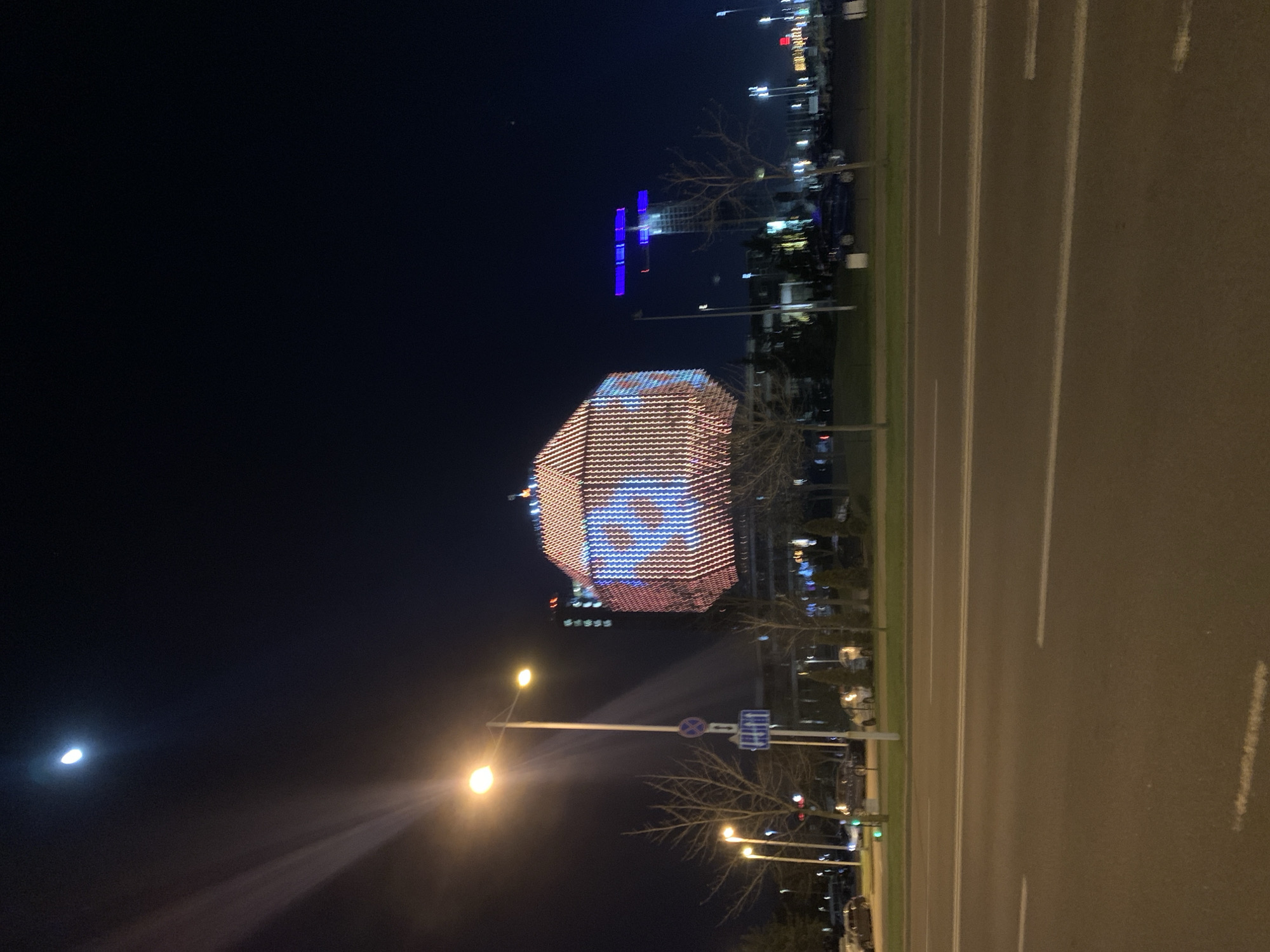 The image shows a nighttime scene in Minsk, Belarus, featuring the National Library of Belarus. The library is illuminated with a pattern of lights, primarily in shades of pink and blue. In the distance, another building is lit up in blue. A street runs along the right side of the frame, with streetlights illuminating the scene and creating light flares. The sky is dark, with a crescent moon visible. Trees are visible along the roadside, some of them bare. Street signs are visible. There are cars parked along the street.