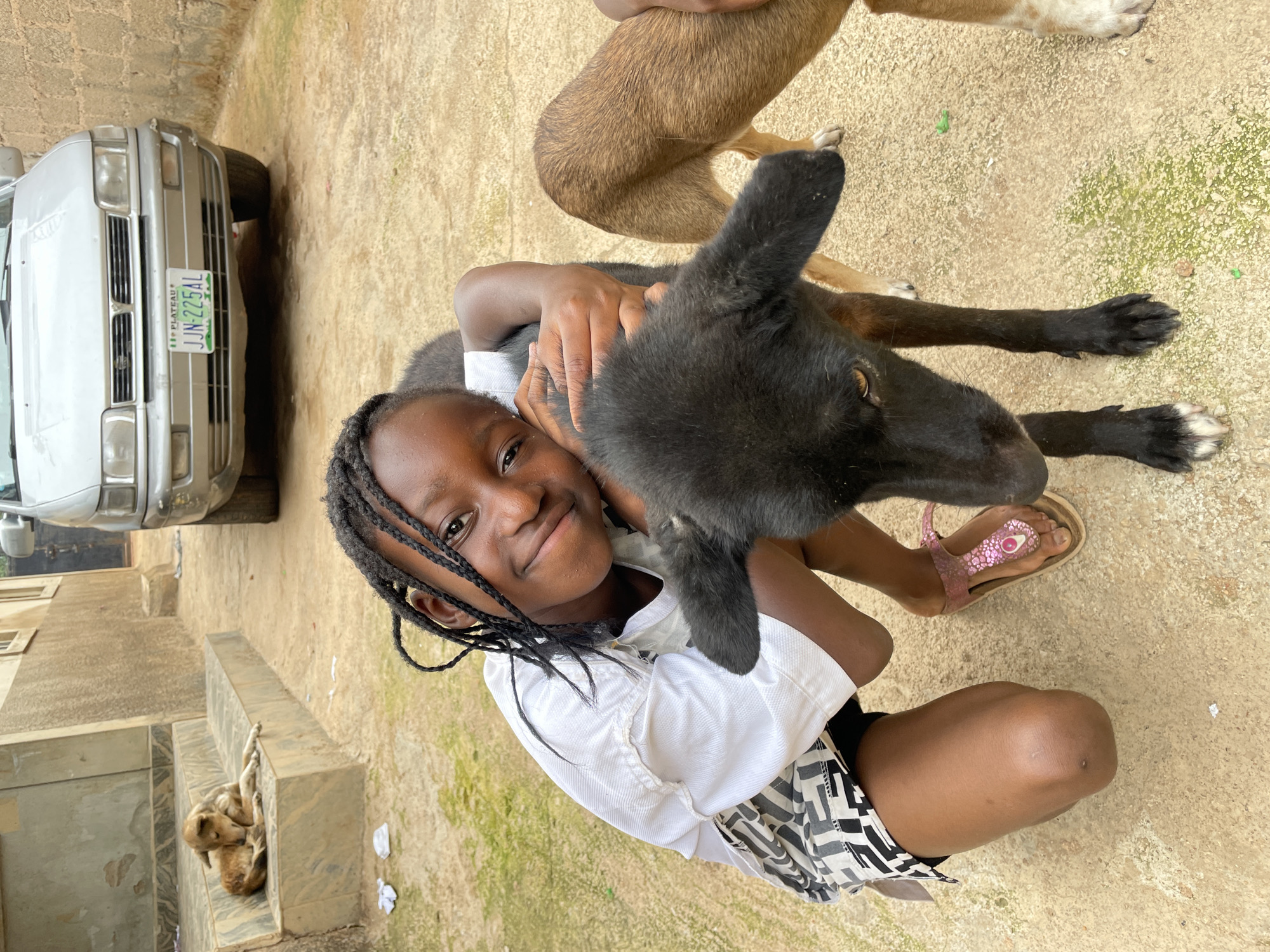 In Gura Top, Nigeria, a young Black girl with braided hair smiles at the camera while kneeling and hugging a black puppy. She wears a white t-shirt and patterned shorts, along with pink sandals. The puppy is black with brown undertones visible around its eyes and ears. In the background, a tan dog rests on a set of stone stairs. A silver car with a Nigerian license plate is parked in front of a brick wall. The ground is a mix of concrete and dirt, with scattered greenery.