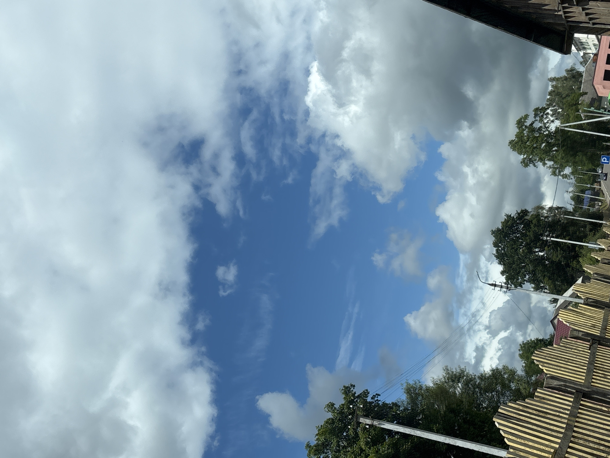 The image depicts a cloudy sky over a rural or suburban area, likely in or near Капыльскі сельскі Савет, Belarus, based on the provided context. The sky is a mix of bright blue patches and large, white and gray cumulus clouds. In the foreground, a wooden fence with yellow and white vertical slats runs diagonally across the lower left portion of the frame. To the left of the fence, a tall, light-colored pole, possibly a utility pole, is visible with wires stretching across the frame. Green trees are scattered throughout the scene, with a denser cluster on the left side of the image. A streetlamp is visible behind the fence, with wires running from the utility pole. In the background, several buildings with reddish roofs and some taller, modern structures are partially obscured by trees and clouds. Cars are visible on a road in the distance. The overall impression is of a typical day with variable cloud cover in a Belarusian landscape.