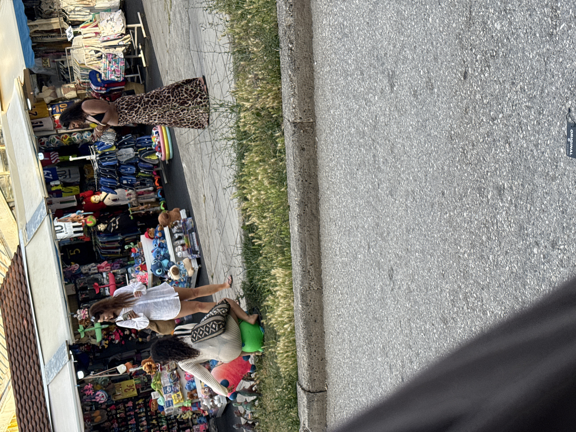 The image captures an outdoor market scene, likely in Budva, Montenegro, as indicated by the context. The photograph is oriented vertically, showing a bright, sunny daytime setting.
In the foreground, a gray asphalt road or path dominates the right side of the frame, partially obscured by a dark shadow in the bottom right corner. A concrete curb separates the asphalt from a strip of dry, light green grass and weeds that runs horizontally across the middle of the image.
On the left side of the frame, behind the grassy strip, is an active market stall. A white awning covers the stall, which is brimming with merchandise. Three women are visible, browsing the items. One woman with dark, curly hair wears a black top and a long, flowing leopard-print skirt; she is positioned in the upper part of the stall area, appearing to examine items on a rack. Below her, another woman with long, dark hair, dressed in a white, loose-fitting long-sleeved shirt and light-colored shorts, also looks through the displayed goods. Partially visible behind her, a third woman with dark, curly hair wears a white top with a black geometric pattern and carries a patterned shoulder bag.
The market stall itself features a diverse array of products, including clothing such as football jerseys with numbers like "10" and "30," colorful bags, and numerous stuffed toys, including blue characters, a green plush, and a red blob-like item. The merchandise is arranged on racks and shelves, extending to the ground level.
A notable detail is a black tag with the word "ORIGINAL" printed in white, visible on the asphalt in the mid-right section of the image. The overall atmosphere suggests a casual shopping experience at a bustling tourist market.