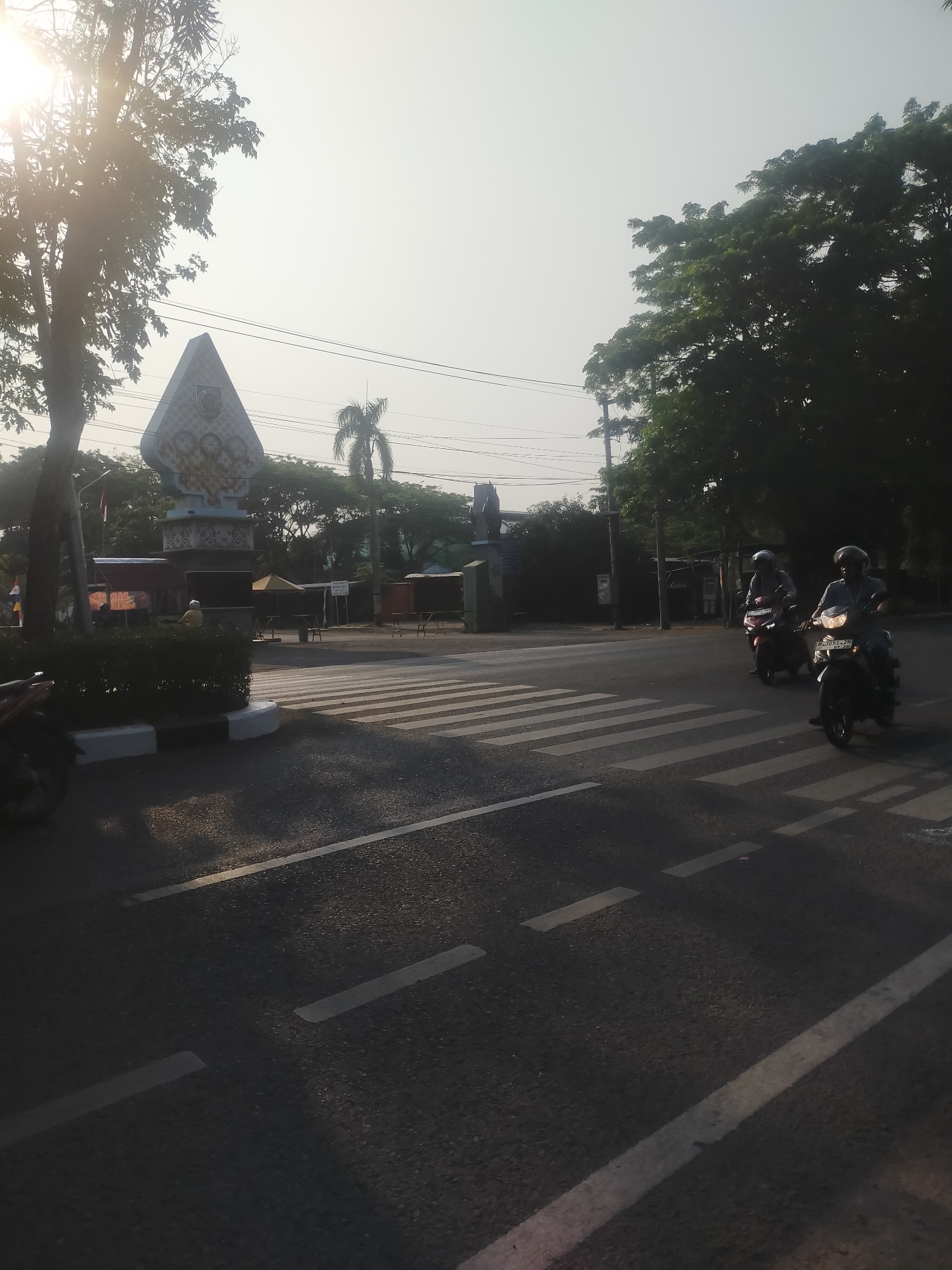 The image depicts a street scene at what appears to be a crosswalk in Pedagangan, Indonesia. The sun is low in the sky, casting long shadows and creating a bright, hazy atmosphere. In the foreground, a crosswalk with white stripes is visible on the asphalt road. Two individuals are riding motorcycles, one in front of the other, heading towards the viewer. Both riders are wearing helmets. The motorcycle in the foreground is white and the one behind it is red.
To the left of the road, there is a paved area with a white curb and a hedge. A motorcycle is parked on the left side of the image, partially visible. Beyond the hedge, a triangular monument with intricate carvings and what appears to be artwork is visible. In the background, there are several trees, some of which are large and leafy, and a tall palm tree. Behind the monument and trees, there are various structures that appear to be stalls or small buildings, some with yellow awnings. Overhead, electrical wires are strung across the sky. The overall impression is a somewhat hazy outdoor scene, possibly in the morning or late afternoon, with traffic present at a pedestrian crossing.