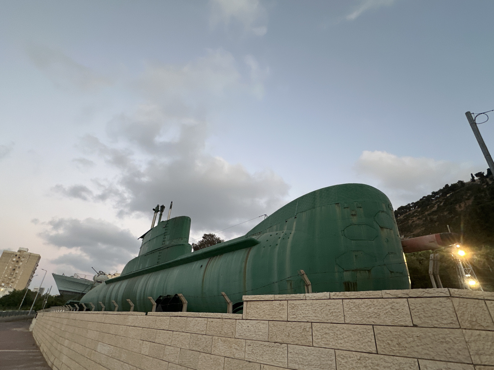 The image captures a large, green submarine displayed outdoors in Haifa, Israel, likely as a museum exhibit. The submarine is the dominant subject, positioned prominently in the foreground, with its hull extending across the frame. A section of a light-colored stone wall runs along the bottom of the image, suggesting a walkway or viewing area. In the background, a modern apartment building and a distant hill with trees can be seen. The sky is overcast with clouds, indicating either early morning or late afternoon light. Another vessel, possibly a ship or another submarine, is partially visible behind the main subject, and a street lamp is visible on the right. There are no people or animals present in the image, and the submarine appears stationary.