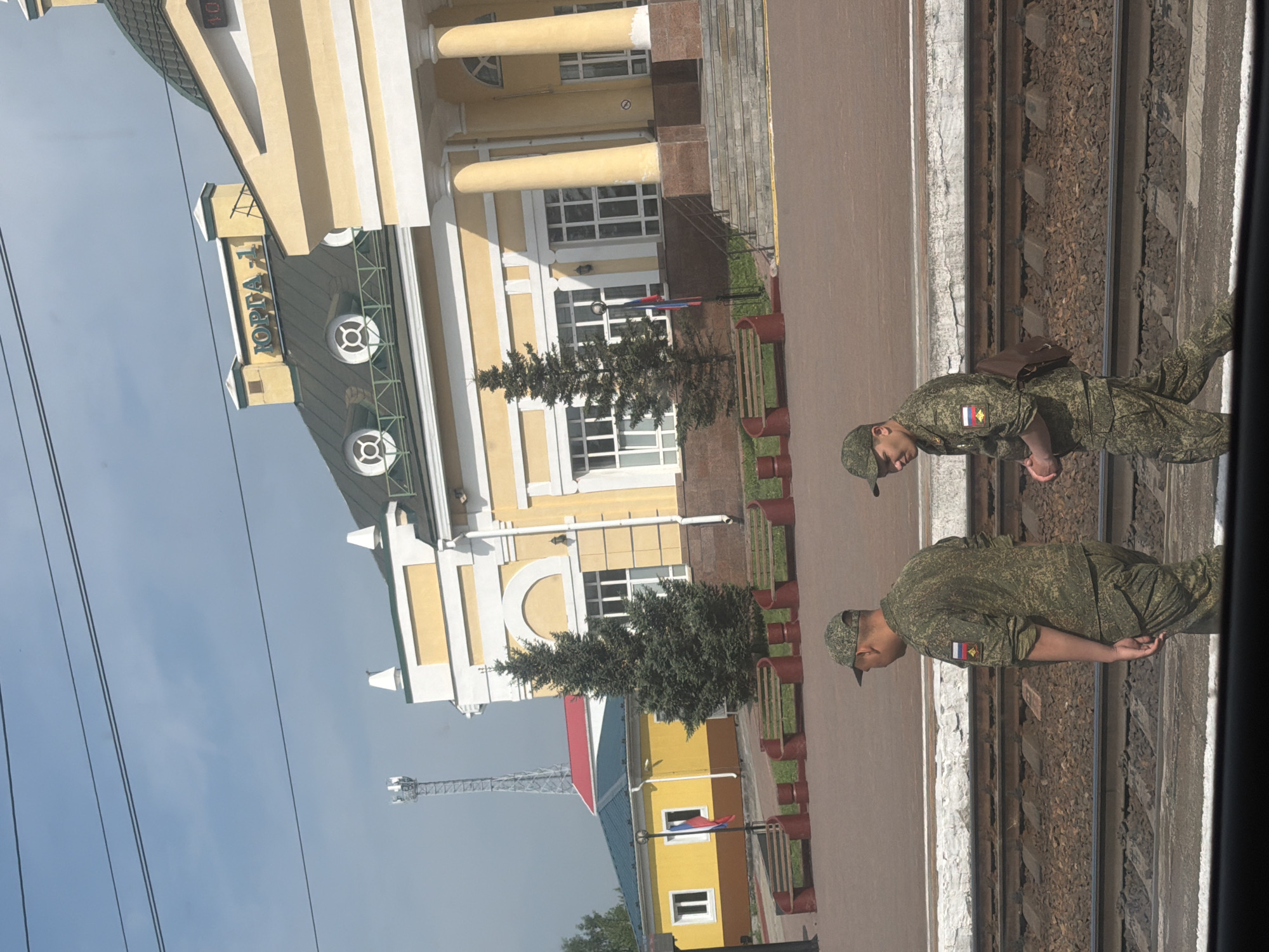 The image shows a train station in Yurga, Russia. The station building is yellow with white trim and a green roof, featuring several arched windows and large columns. The word "ЮРГА-1" (YURGA-1) is visible on the roof of the building. In the foreground, two men in camouflage military uniforms are walking on a paved area next to train tracks. One man, closer to the camera and on the left, is walking away from the viewer, while the other man, on the right, is walking towards the viewer. Both men are wearing caps, and the man on the right has a brown backpack slung over his shoulder. Several evergreen trees are planted in front of the station, and benches are visible along the platform. A cell tower can be seen in the background to the left of the station.
