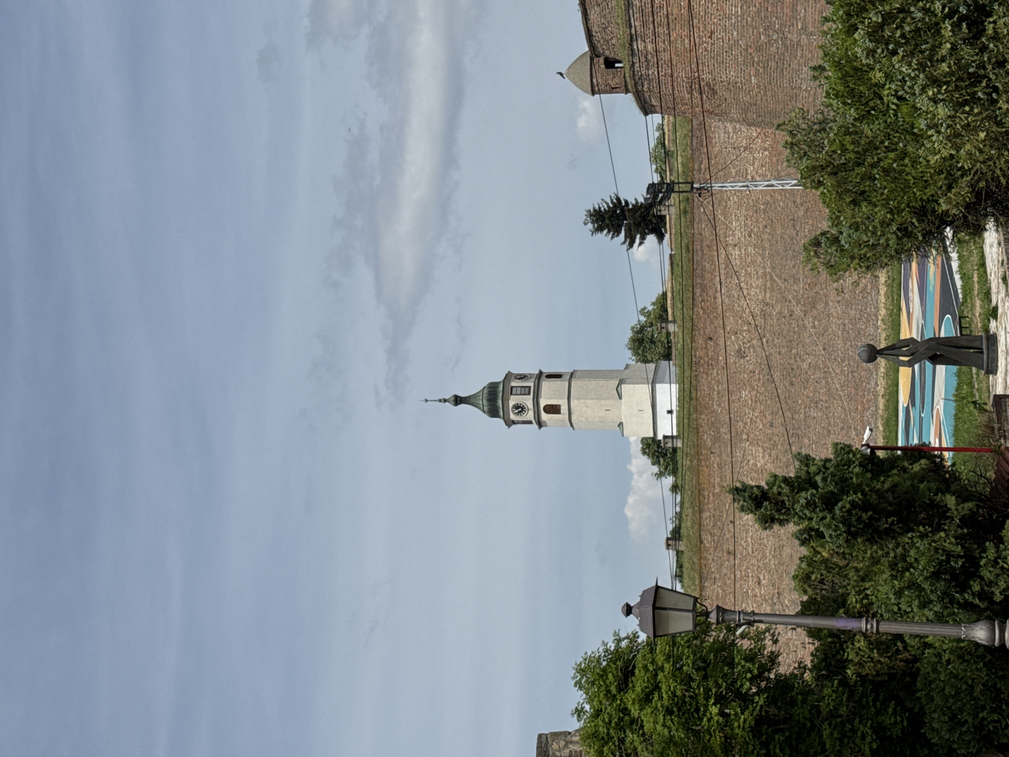 The image shows a view of the Belgrade Fortress, Serbia. The sky is light blue with scattered clouds. In the center of the frame is the clock tower, a landmark of the fortress. A brick wall lines the top right of the image. A grassy area with a few trees and power lines leads towards the tower. A sculpture stands along the right side. In the lower right, there is a street lamp with green foliage around it.