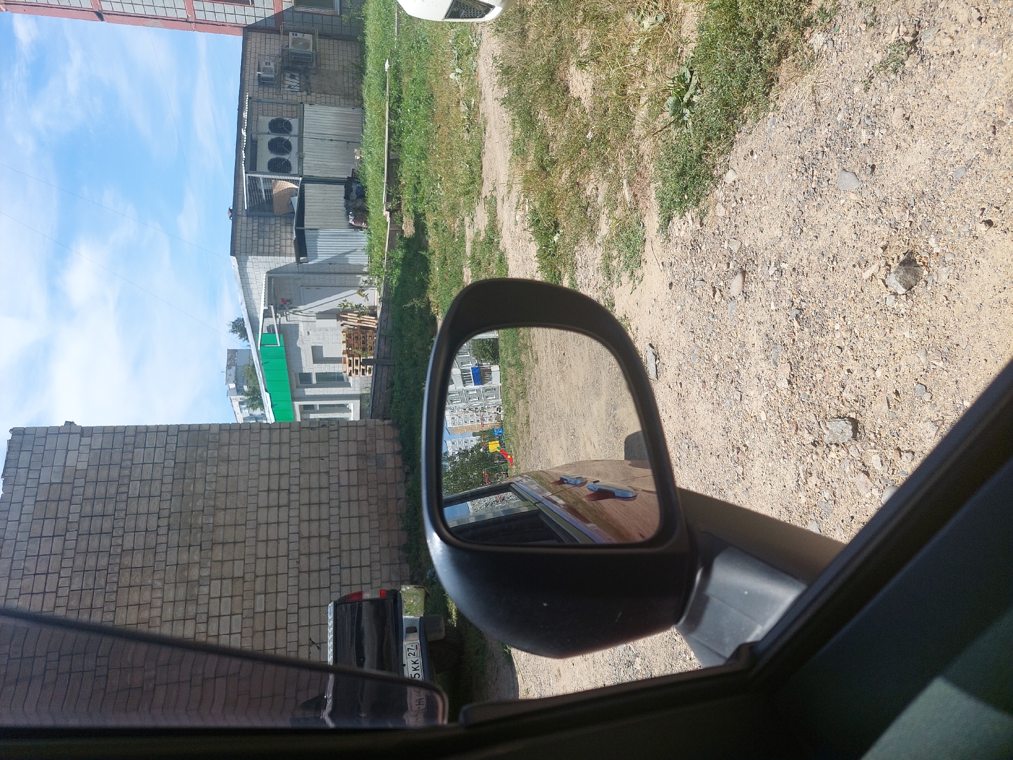 The image shows a view from inside a car in Amursk, Russia, looking out through the driver's side window and side mirror. In the foreground, the interior of the car's doorframe is visible on the left. The side mirror reflects a street with buildings and a playground. Outside the car, a brick building is prominent on the left, with a black pickup truck parked in front of it. Beyond the truck, there are other buildings, including one with green accents and large industrial air conditioning units on its roof. The ground is unpaved and covered with gravel and sparse grass. The sky is blue with some clouds. The license plate of the pickup truck is partially visible, showing Russian characters and numbers.