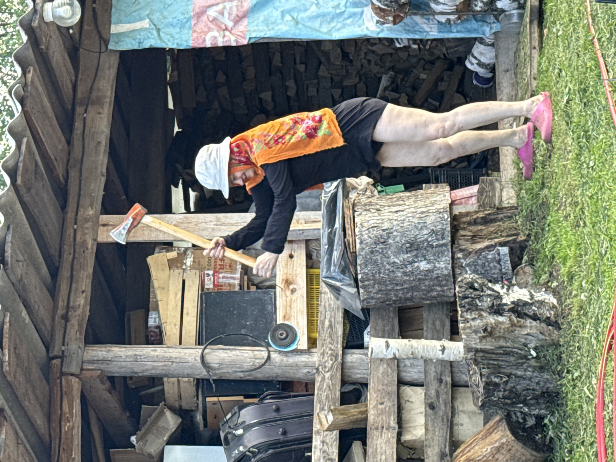 An individual wearing a white hat, an orange and red patterned headscarf, a black long-sleeved shirt, and a dark skirt is holding an axe with both hands, poised as if to swing it. The person is standing on green grass in front of a rustic wooden structure, possibly a shed or outbuilding, which is filled with various items including lumber, boxes, and what appears to be a black suitcase. The background shows a pile of stacked firewood and a blue and pink tarp hanging from the structure. The overall setting suggests a rural or outdoor environment. The location is identified as Rosstani, Russia.