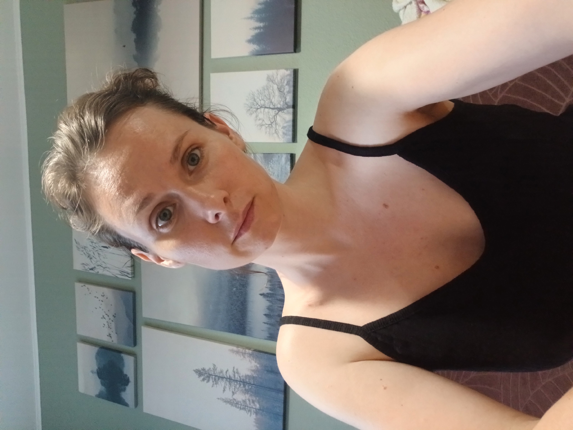 A selfie taken from a slightly high angle shows a woman with light skin and light brown hair, pulled back from her face, looking directly at the camera. She has blue eyes and a neutral expression. She is wearing a black tank top with thin straps. Behind her, the wall is painted a muted green. Hanging on the wall are several framed landscape artworks depicting trees, fog, and bodies of water in a minimalist, blue-toned style. The overall impression is of a domestic indoor setting, possibly a bedroom or living room. No specific location in Yekaterinburg, Russia can be determined from the image.