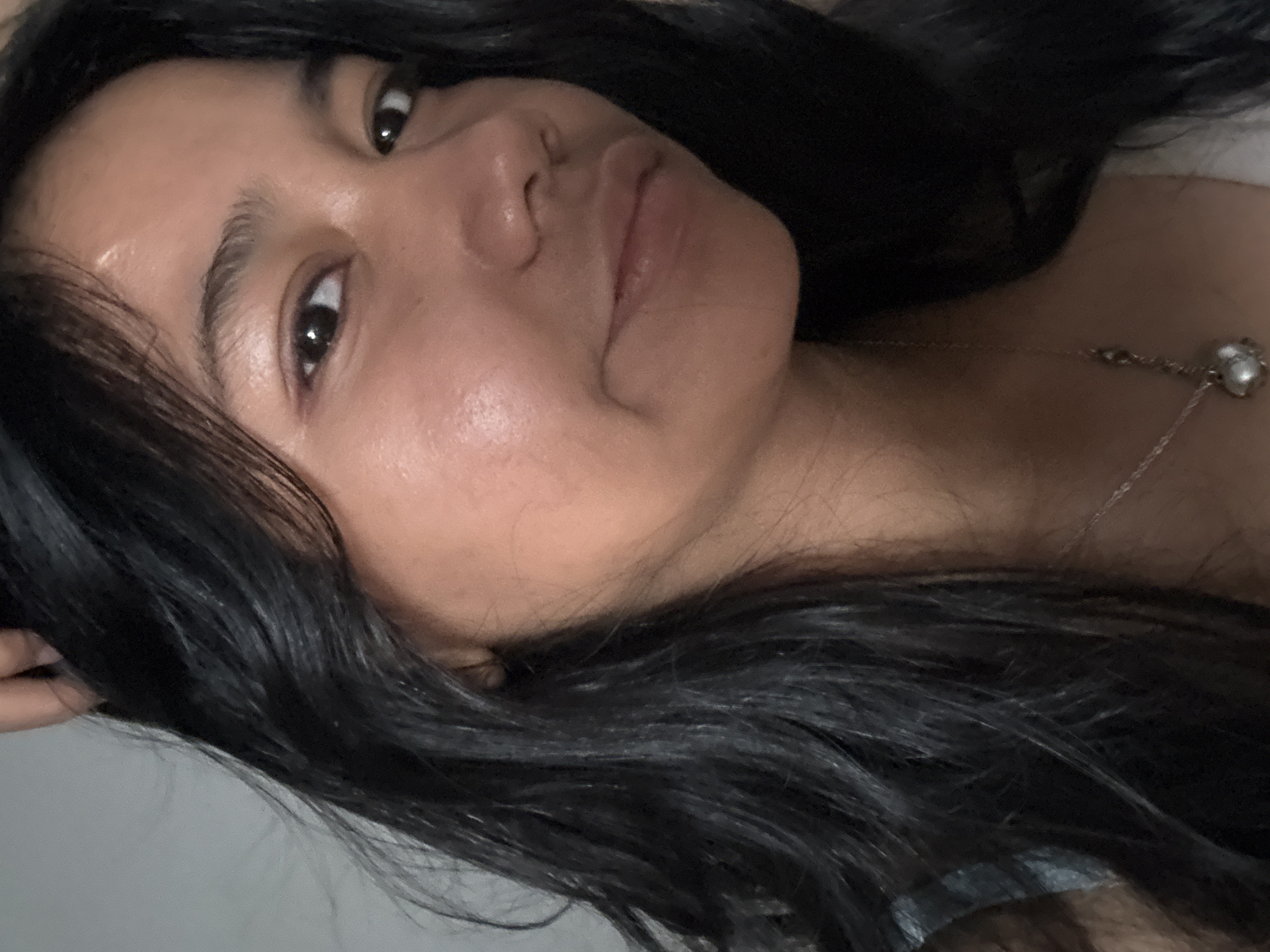 A close-up, self-portrait style photograph shows a young woman with long, dark, wavy hair that partially obscures her face. She is looking towards the camera with a neutral expression. Her skin appears slightly dewy, and she is wearing a delicate silver necklace with a pearl pendant. The background is a plain, light-colored wall, suggesting an indoor setting. The image does not contain any information about a specific location in North Jakarta, Indonesia, or any actions or interactions.