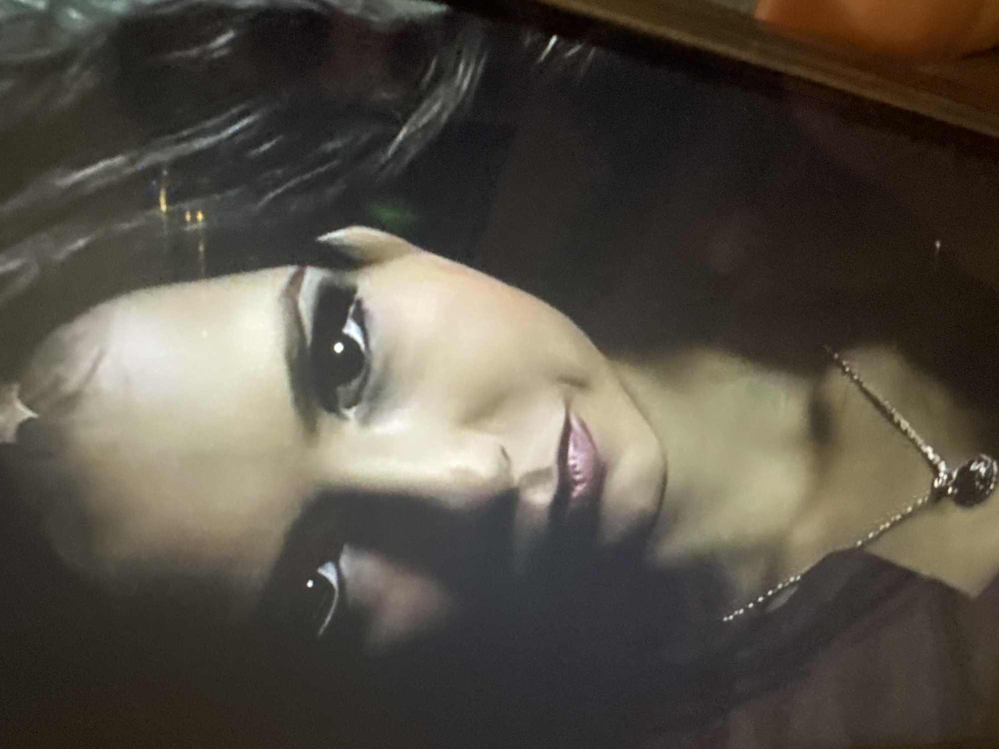 The media file presents a close-up portrait of a person, likely a woman, captured in a dimly lit setting. The subject's face is prominent, oriented diagonally from the upper left to the lower right of the frame. She has dark hair, which is visible across the top and left side of her head. Her eyes are dark and open, looking directly at the viewer, and her lips are slightly parted in a subtle smile or smirk, accentuated by a pinkish-red hue.
She is wearing a silver-toned necklace with a dark, round pendant that rests on her chest, visible towards the lower right. The overall lighting is low, casting shadows that highlight the contours of her face and create a soft, somewhat mysterious atmosphere. The background is largely dark and indistinct, offering no specific location cues. The image appears to be a photograph of a screen, contributing to a slightly grainy texture. No discernible activity is taking place beyond the subject looking at the camera. There is no visible text.