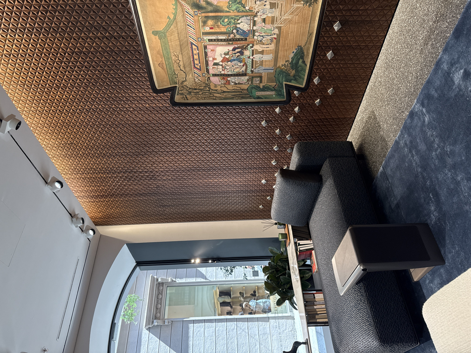 This media file captures a stylish interior space, likely a lounge, showroom, or upscale living room, characterized by modern design and unique decorative elements. The room appears brightly lit, suggesting it is daytime.
The most striking feature is a large, intricately patterned accent wall or ceiling. This surface is covered in dark brown, small, three-dimensional geometric shapes, resembling pyramids or facets, creating a textured, almost sculptural effect. Mounted on this patterned surface is a prominent, ornate, traditional Asian-style painting, possibly Chinese or Japanese, framed in black. The artwork depicts multiple figures in an elaborate setting with classical architecture and lush gardens. Scattered across the patterned wall, seemingly at random, are numerous small, silver-colored pyramidal studs, adding a futuristic contrast to the traditional artwork.
In the foreground, a modern, dark grey upholstered sofa with a textured fabric sits on a plush, deep blue rug. An integrated or attached black side table is positioned over the armrest of the sofa. Adjacent to the blue rug, a lighter grey, possibly stone-textured floor or rug, is visible.
To the left, a clean, white wall features modern track lighting with multiple adjustable spotlights, illuminating the space. This wall curves gracefully into an arched opening, revealing a large window or doorway. Through this opening, an exterior scene is visible, showing parts of a building with white panels and some green foliage. Several blurred figures, presumably people walking, can be seen outside, indicating a street or pedestrian area. On a low surface near the window, a green potted plant and a stack of books are discernible.
The overall atmosphere is sophisticated and curated, reflecting a blend of traditional art and contemporary design, consistent with a high-end setting in a city like Milan, Italy. No legible text is visible in the image.