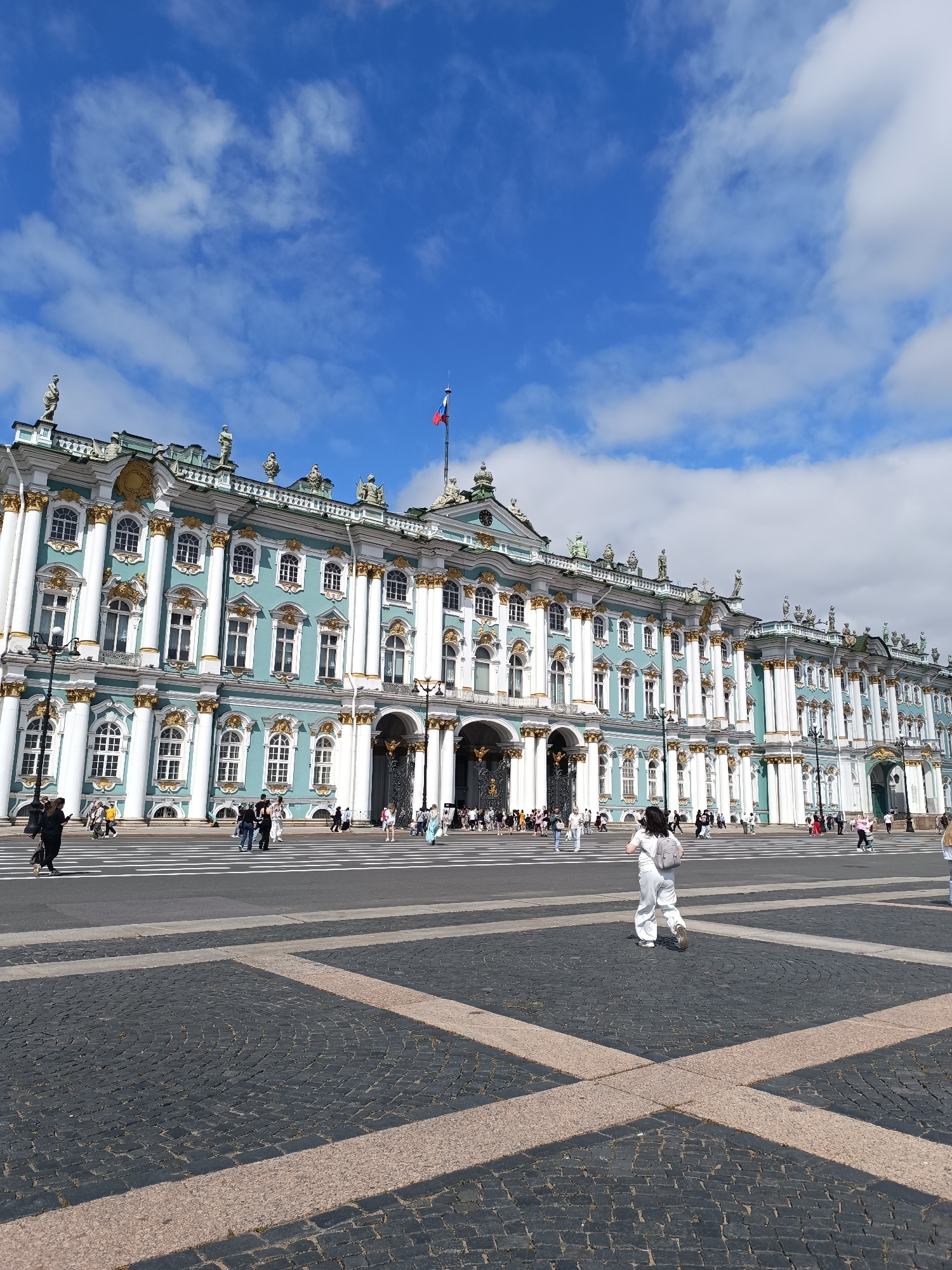 The image shows a wide-angle view of the Winter Palace, a prominent building in Saint Petersburg, Russia, situated on Palace Square. The palace is a grand structure with light blue facades, white columns, and ornate gold detailing, characteristic of Baroque architecture. It is topped with numerous statues and decorative elements. A Russian flag flies from a flagpole atop the building.
In front of the palace is Palace Square, paved with dark cobblestones with lighter rectangular inlays. A pedestrian crossing marked with white stripes is visible on the square, leading towards the palace entrance.
Several people are scattered across the square. In the foreground, a person wearing white clothing and a backpack is walking away from the camera, crossing the paved square. Further back, groups of tourists are seen walking and standing, some near the zebra crossing and others in front of the palace. The sky is blue with scattered white clouds. The overall scene depicts a sunny day in a public, iconic urban location.