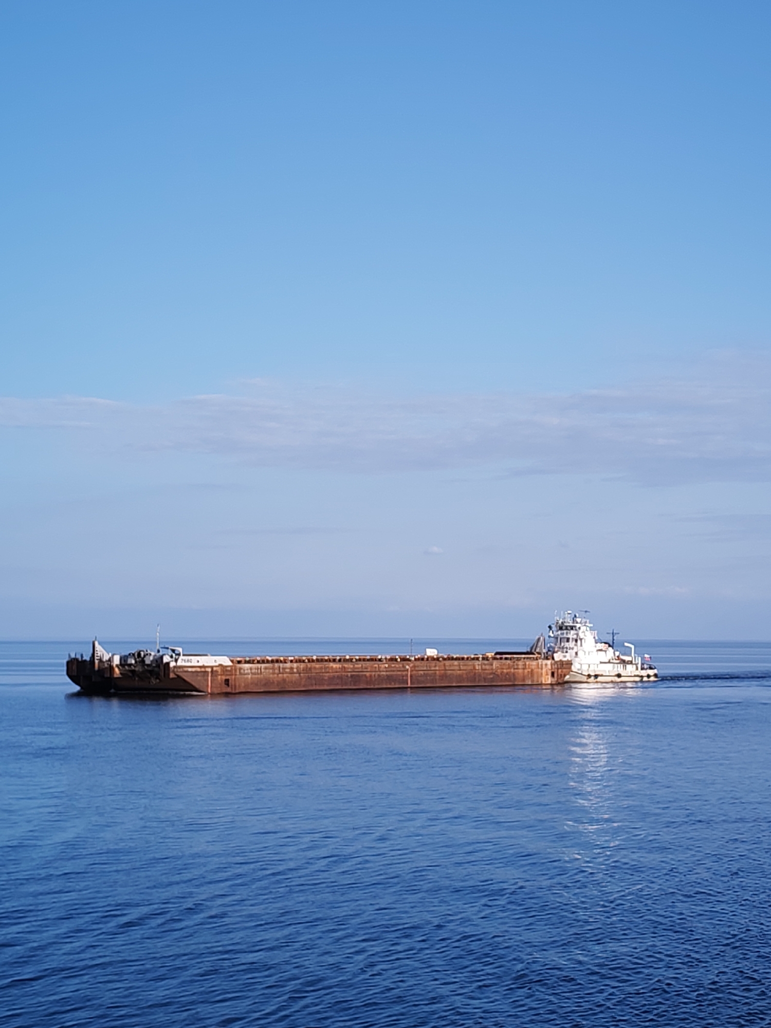 A long, rust-colored barge is being pushed across a body of water by a white tugboat. The sky above is clear with a few scattered clouds. The water is deep blue, with slight ripples on the surface. The location is unknown.