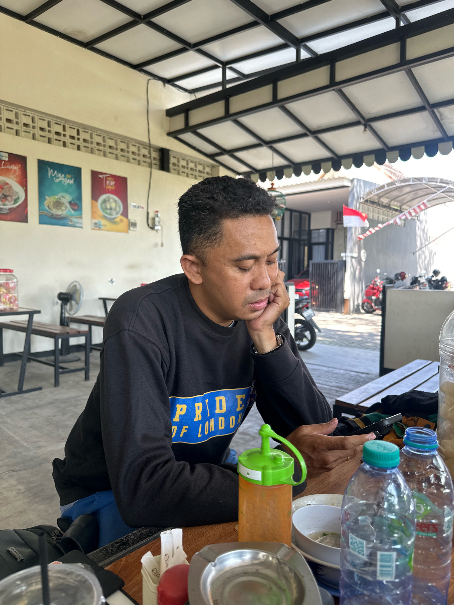 A man with short, dark hair sits at a wooden table outdoors, resting his head on his hand and looking down. He wears a black sweatshirt with blue and yellow lettering that reads "PRIDE OF LONDON". In front of him are several objects: a bottle of orange sauce with a green lid, a metal ashtray, a white bowl with food, and two plastic water bottles, one blue and one clear. Behind him, a ceiling fan is mounted on a wall, and a jar with red items sits on a shelf. Posters depicting food are visible on the wall. The background shows a building with a red and white Indonesian flag displayed and several motorcycles parked outside. The scene appears to be a casual dining or resting area, possibly in Mojokerto, Indonesia, given the Indonesian flag and common Indonesian food items visible.
