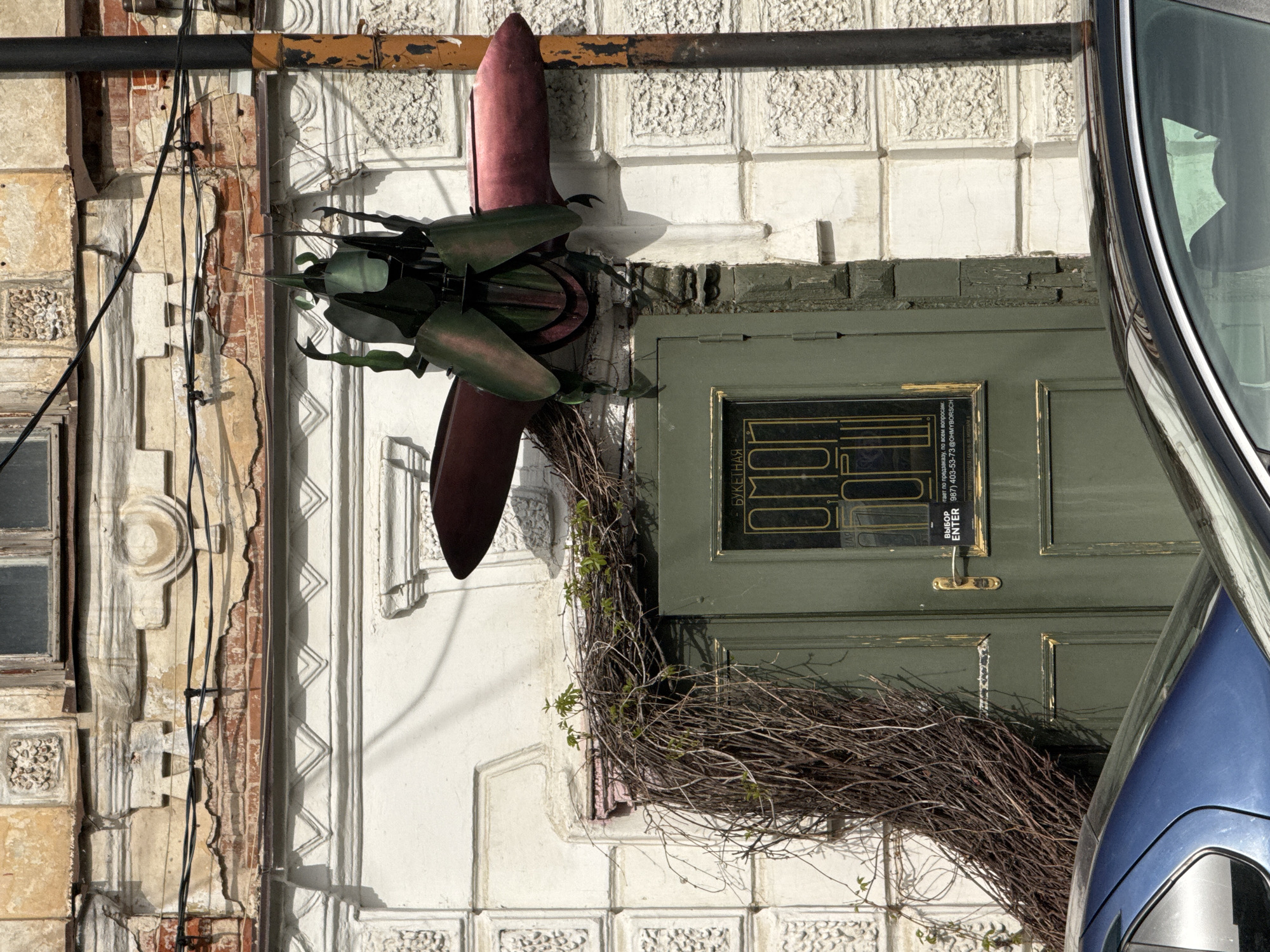 The image captures a building exterior in Kazan, Russia, evidenced by Cyrillic text on the door. The building features a white façade with textured square blocks. A green wooden door is adorned with a decorative wreath of twigs and some sparse greenery. Above the door, a maroon-colored, propeller-shaped decoration is mounted. A black metal pipe runs horizontally across the upper part of the frame, with some rusted areas. To the right, the corner of a blue vehicle is visible, possibly a car. The left side of the image shows a section of an older building with crumbling plaster and exposed brick. Black electrical wires are also visible against this part of the façade.