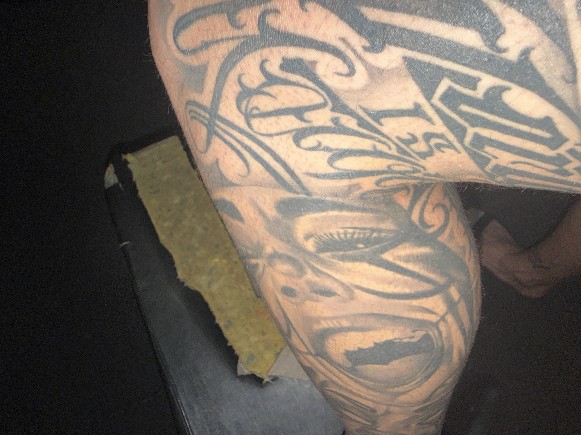 An individual's tattooed leg is visible, with a large, dark tattoo covering most of the skin. The tattoo features stylized lettering and abstract designs. The leg is positioned near a dark surface, possibly a car interior, with a torn piece of yellow padding visible in the background. The lighting is dim, casting shadows on the scene. The individual's foot is also partially visible at the bottom of the frame.