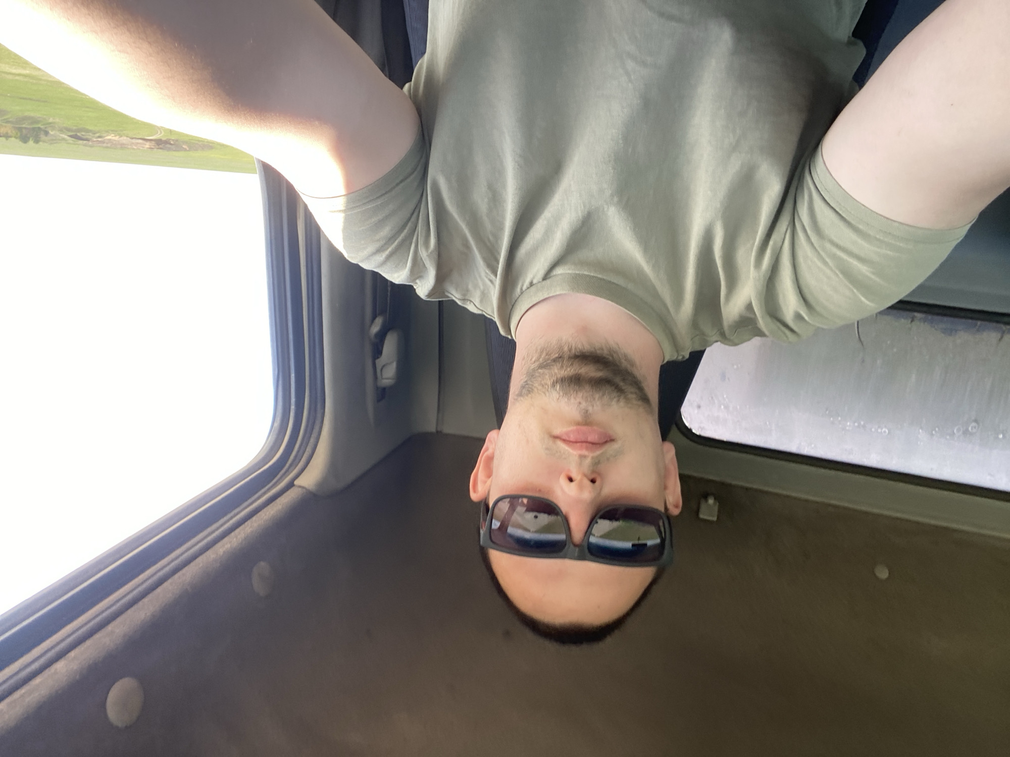 The image shows a man inside what appears to be a vehicle. He is wearing sunglasses and an olive-green shirt. The picture is taken from an unusual angle, making him appear upside down. Visible through the vehicle's windows is a landscape featuring a field. There is no evidence to link the image to Kapsal, Russia.