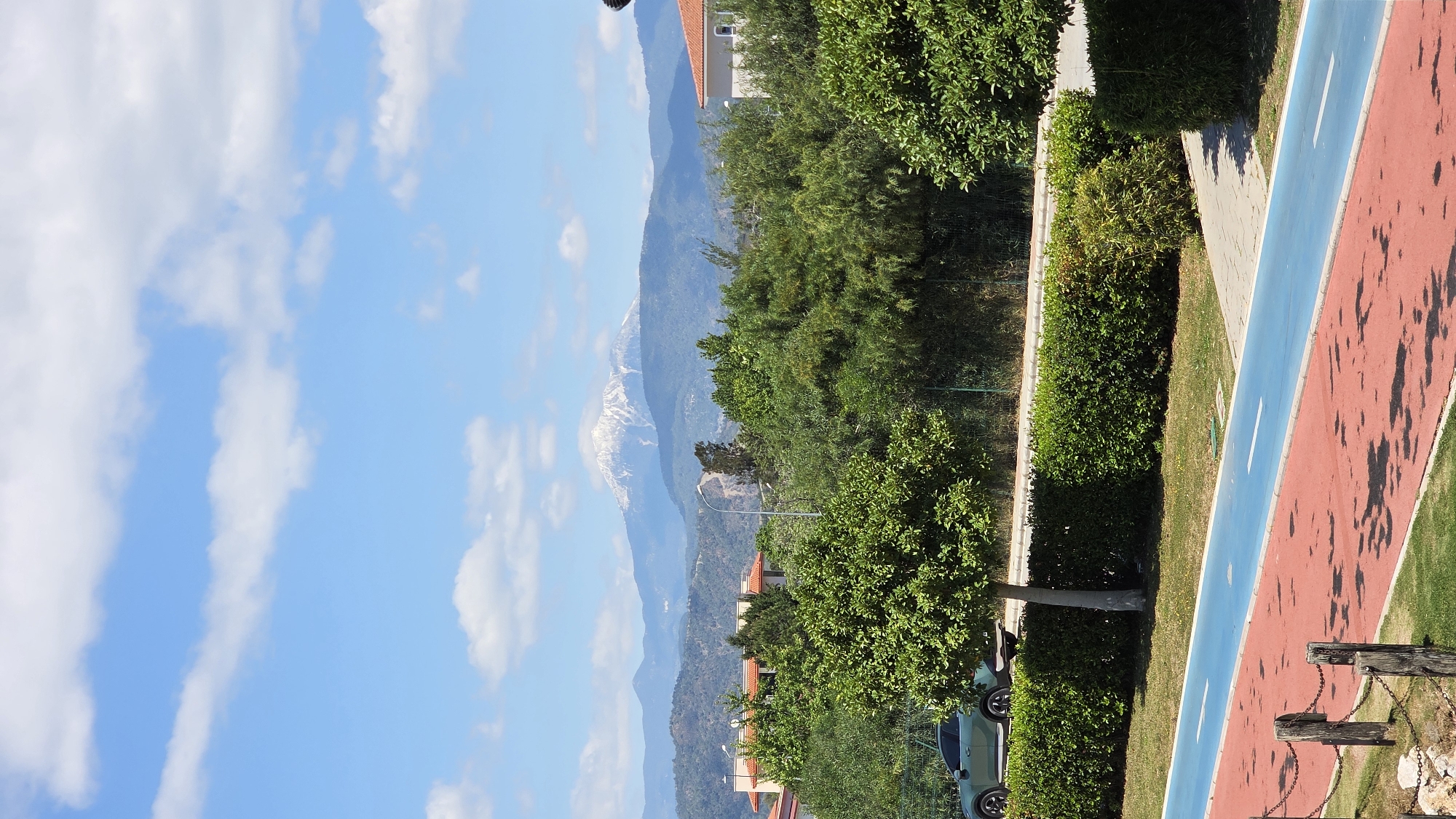 A sideways, elevated daytime view shows a red and blue walking/jogging path flanked by green foliage in Fethiye, Turkey, with a snow-capped mountain range in the distance. The sky is blue with scattered white clouds. A dark green car is parked near a tree. A low, decorative fence made of posts and chains is in the foreground. The location is likely near the beach and the mountains are visible in the distance. Some buildings are seen further back.