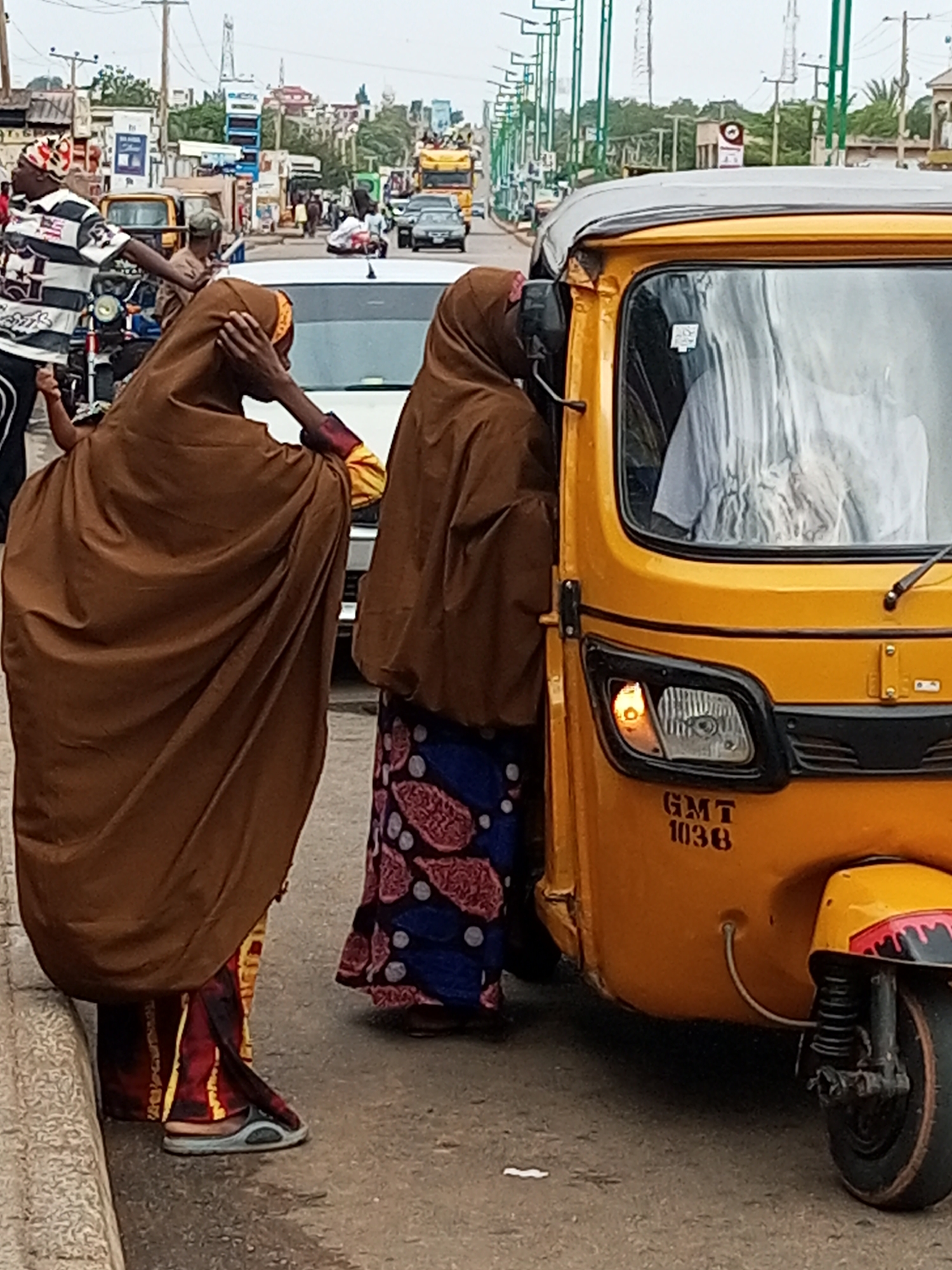 Two women, dressed in traditional Islamic attire, stand near a yellow auto-rickshaw on a street. The woman on the left wears a brown hijab and a colorful patterned skirt, with a glimpse of a yellow and red garment underneath. She is adjusting her hijab with one hand. The woman on the right also wears a brown hijab and a blue and pink patterned skirt.
The auto-rickshaw is the dominant object in the foreground, with its yellow body and black trim. The vehicle has a visible headlight and a license plate with the text "GMT 1038" in dark lettering. Inside the windshield, a person in a light-colored shirt can be partially seen.
The scene depicts a street in Tempure, Nigeria, with a line of traffic visible in the background, including cars and other vehicles. Buildings and shops line the street, and utility poles with wires crisscross the sky. The weather appears overcast, suggesting a cloudy day. The overall atmosphere seems to be one of a typical urban environment with people engaged in daily activities.