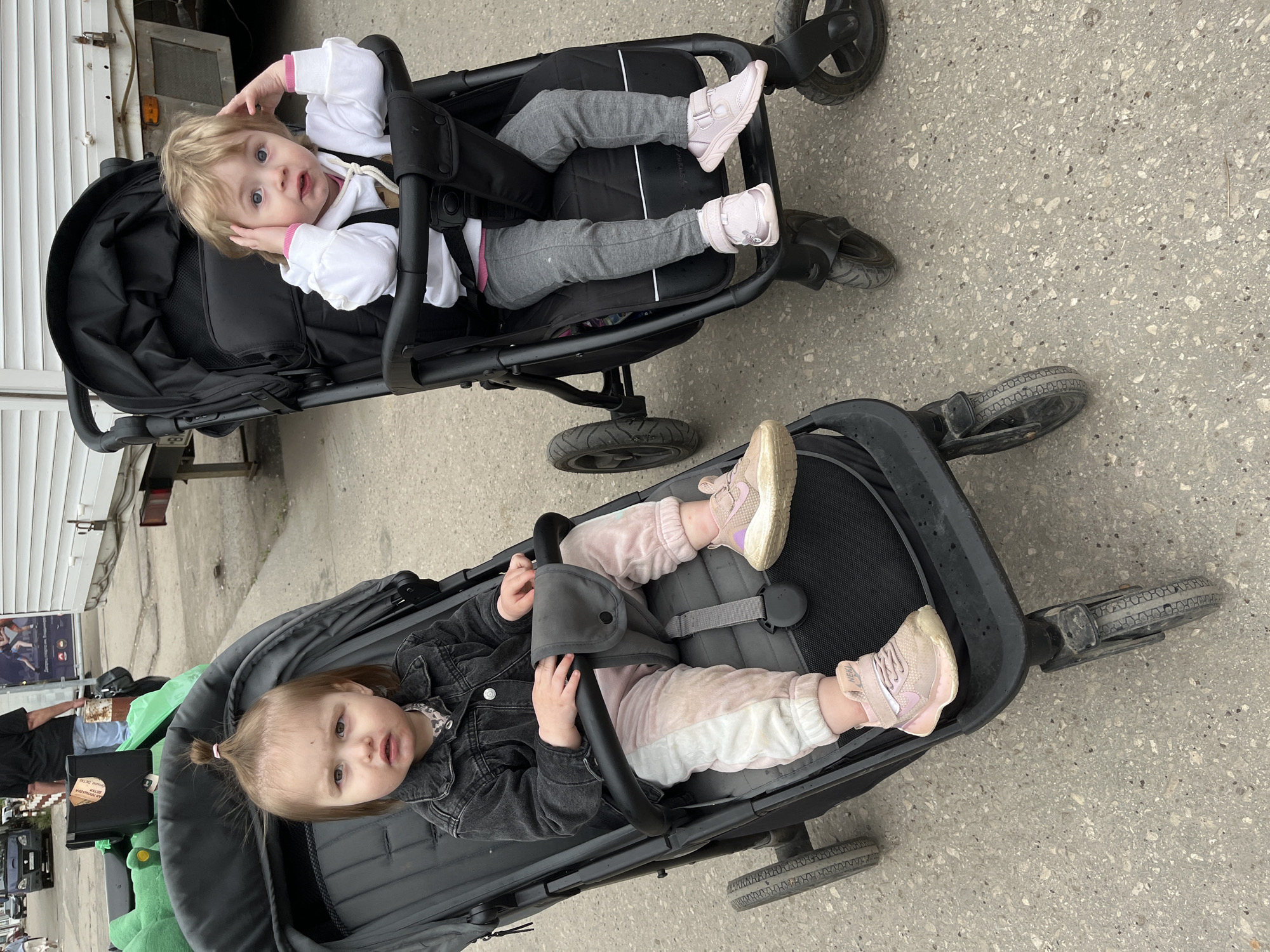Two children are positioned in black strollers on a concrete surface. The first child, located in the top stroller, has fair hair and is wearing a white top, grey trousers, and light pink shoes. The child's hands are near their face. The second child, in the bottom stroller, has brown hair tied in a small ponytail, is wearing a black jacket, pink and white trousers, and pink shoes. The surrounding area shows a street scene; items such as green bags, a black box, and a poster are visible, and part of a vehicle and a building are also within the frame. The location is Novokuybyshevsk, Russia.