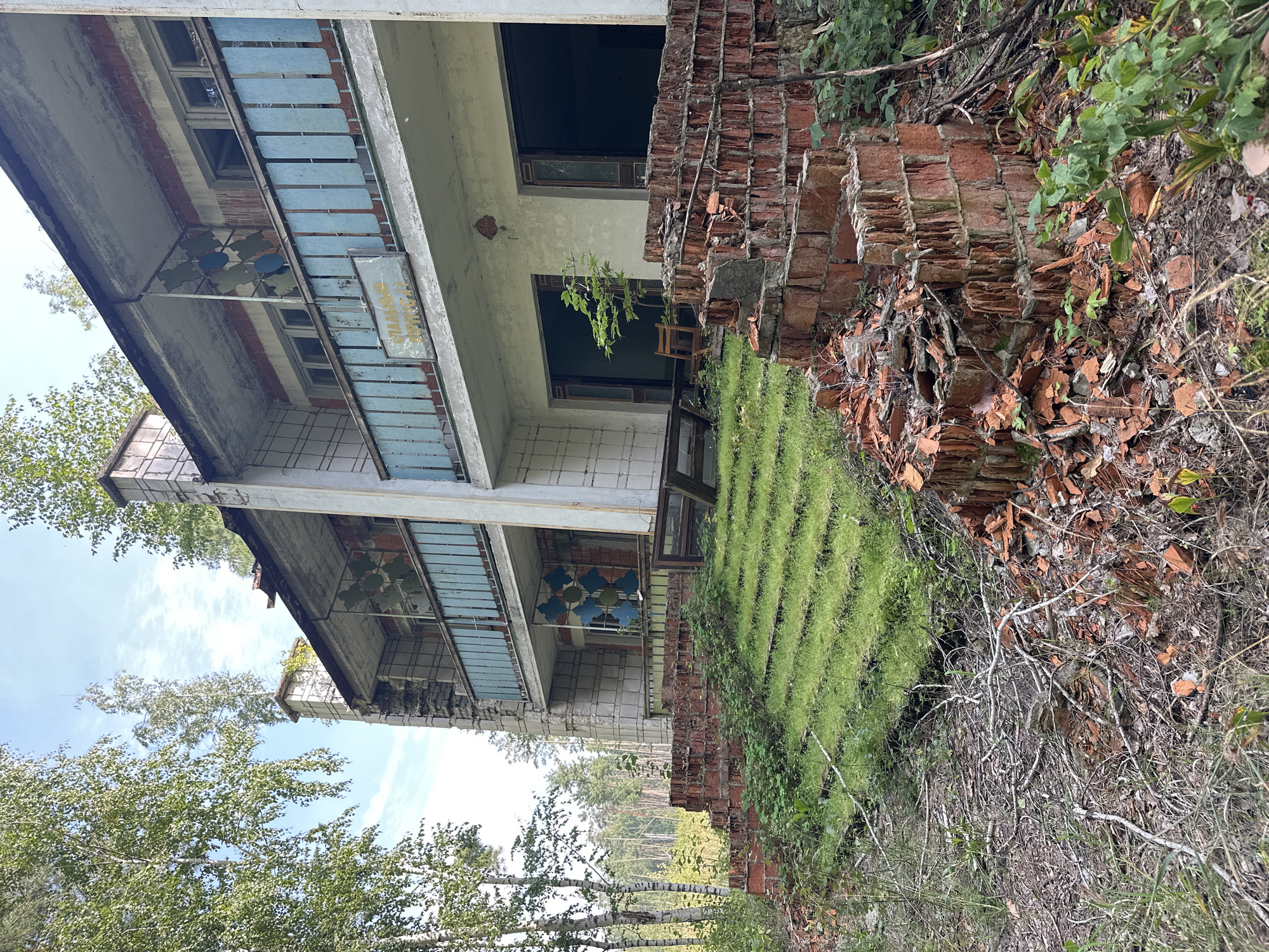 The image depicts an abandoned building with a crumbling brick staircase in the foreground. The building, likely a former residential or communal structure, shows signs of severe neglect, with broken windows, peeling paint, and overgrown vegetation. The staircase is made of red brick, and many of the bricks are broken or dislodged, with grass growing through the gaps. The building itself has multiple stories with balconies, some of which have blue metal railings. A sign with Cyrillic text is visible on one of the balconies. The surrounding environment is overgrown with trees and underbrush, suggesting a rural or natural setting. The sky is visible above, with some clouds and branches of trees. The overall impression is one of decay and abandonment. The location is identified as Sadovka, Russia.