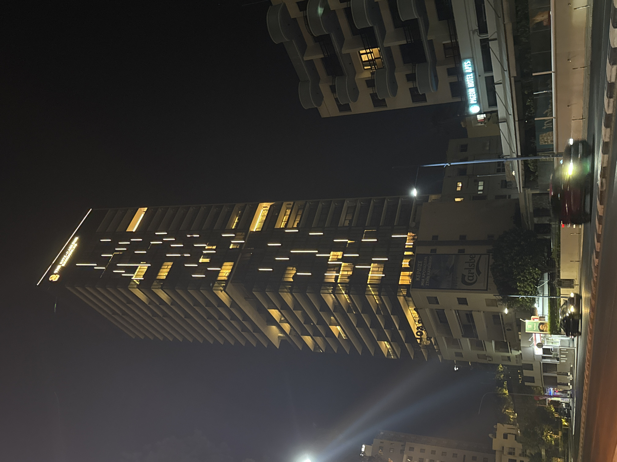 This image shows a nighttime view of a tall, modern skyscraper in Mouttagiaka Community, Cyprus. The skyscraper is illuminated with vertical strips of light that highlight its many floors and balconies. The building's facade is dark, with rows of balconies protruding outwards. At the very top, a sign reads "THE RITZ-CARLTON."
To the right of the skyscraper, another, shorter building with a rounded balcony design is visible. This building also has illuminated windows. Below the skyscraper, there is a lower-rise building with a prominent advertisement for "Carlsberg" and text in Greek that appears to be advertising a competition. Signs for "PIGEON HOTEL APTS" are also visible on a building further to the right.
The street in the foreground is paved and has crosswalk markings. Several cars are present on the street, with one red car in motion, its headlights illuminated. Streetlights are also visible. The overall environment suggests an urban setting at night.