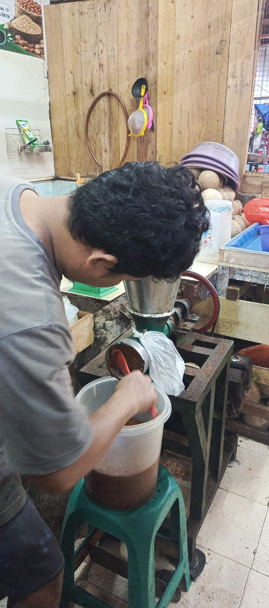 In Sukaresmi, Indonesia, a man with dark, curly hair is shown from the back, bending over a grinding machine. He is wearing a grey t-shirt and dark shorts. The machine, made of metal with a green base, appears to be processing some kind of food product. A red spatula is in his hand, scraping a brown, granular substance from the machine into a translucent plastic bucket. The bucket is resting on a green plastic stool. Behind the man and the machine, a wooden wall is visible, adorned with a circular metal hoop and a colorful hanging object. Various items, including what appear to be coconuts in baskets, buckets, and a red plastic container, are scattered in the background, suggesting a market or processing area. A poster with text is partially visible on the left wall. The floor is tiled.