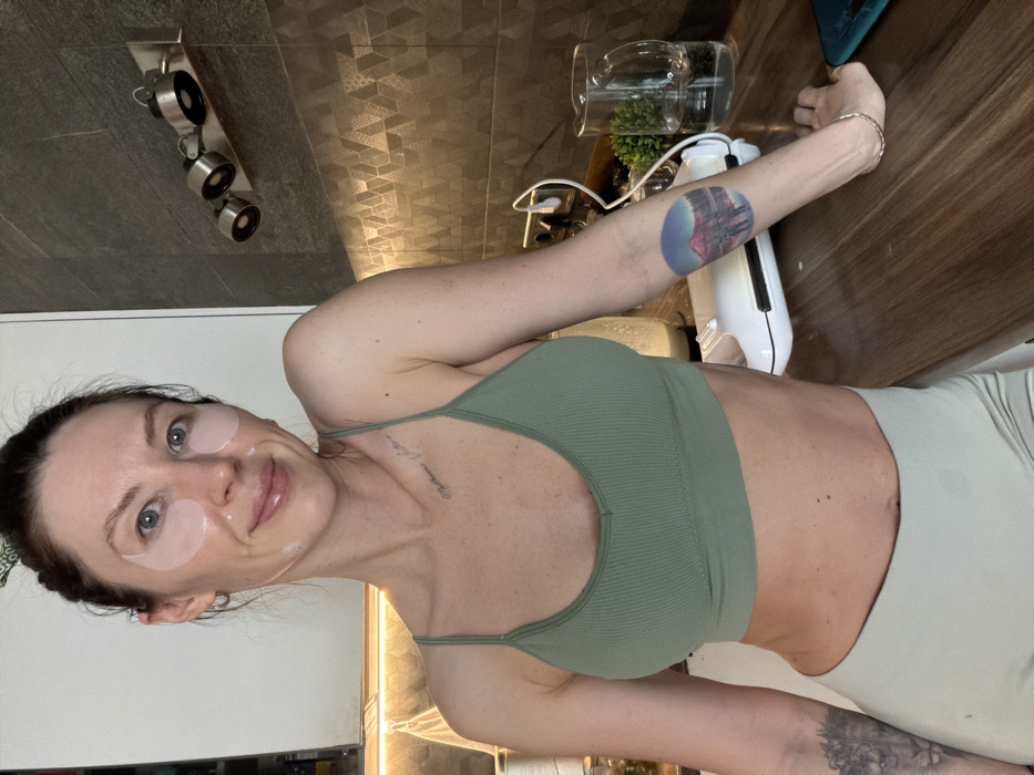 A woman wearing eye patches and a green sports bra poses for a selfie. A tattoo is visible on her arm. Behind her is a countertop with a blender, water pitcher, and plant. A wall with track lighting and a cabinet are also visible. The photograph is rotated to the right.