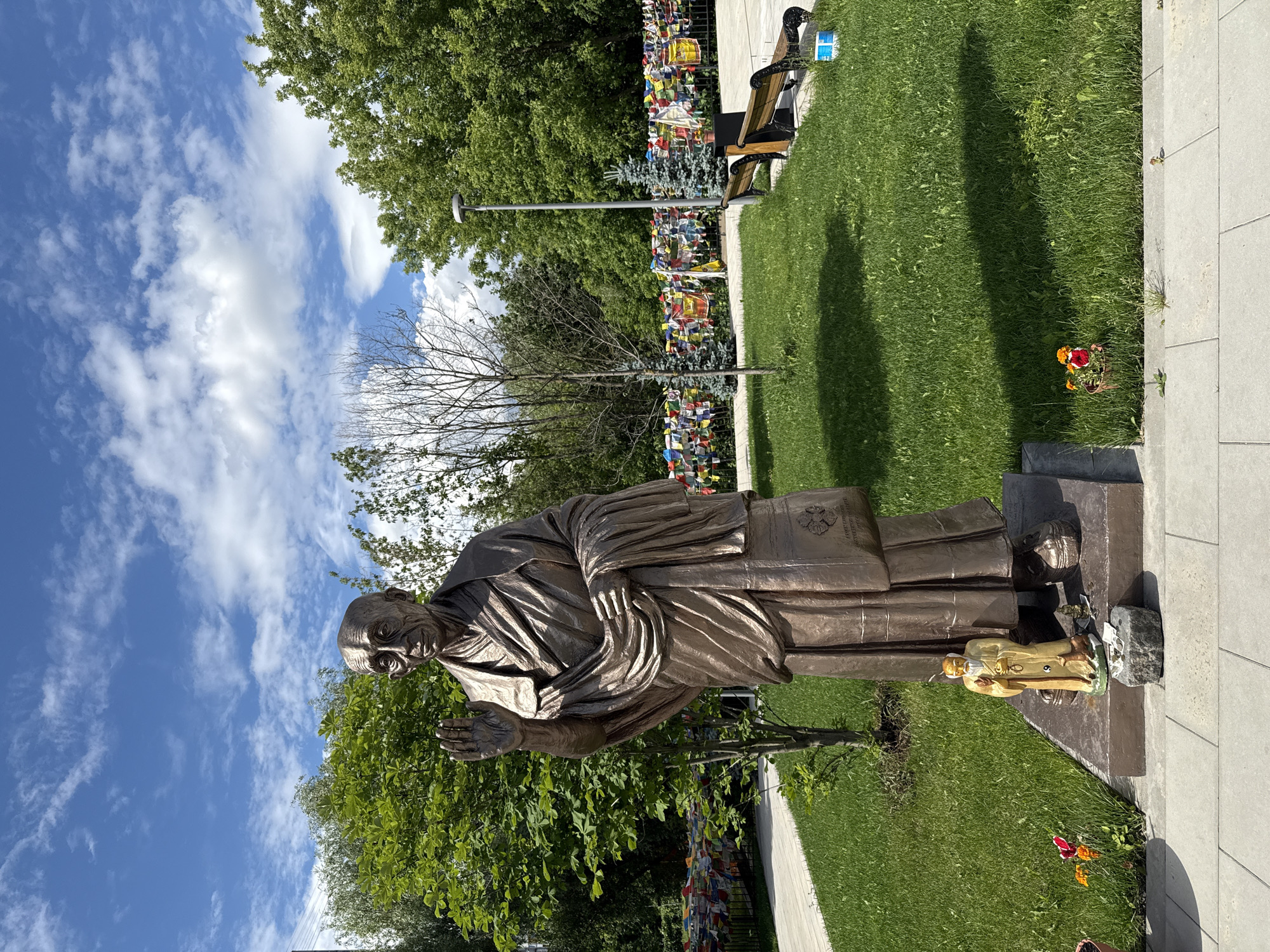 This media file captures a bronze statue of a kneeling figure with hands pressed together in prayer, likely in a public outdoor space. The statue is positioned on a stone base, with a smaller, lighter-colored figurine of a person holding a staff standing at its feet. The background features lush green grass, a paved walkway, and trees. In the distance, a fence is adorned with numerous colorful prayer flags, suggesting a spiritual or commemorative site. A wooden bench is visible to the left of the prayer flags, and a lamppost stands in the mid-ground. The sky above is a vibrant blue with scattered white clouds, indicating fair weather. The lighting suggests it is daytime, possibly late morning or early afternoon, given the shadows cast by the statue and trees. The overall scene conveys a sense of peace and reverence. No discernible text is visible on the statue or surrounding elements.