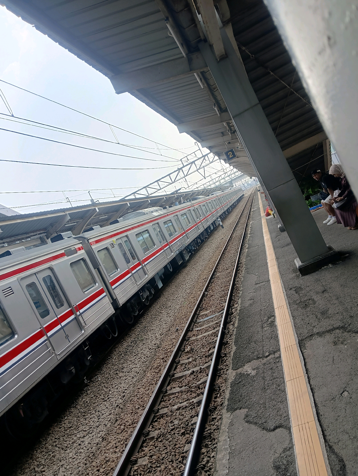 The image depicts the train station in Parung Panjang, Indonesia, featuring a silver train with red and blue stripes parked alongside the platform. The train appears to be a commuter or electric multiple unit (EMU) type. The platform has a textured, gray surface with a yellow tactile warning strip near the edge. Overhead, a roofed structure supported by thick gray pillars provides shelter. In the background, intricate overhead electrical wires and support structures for the railway are visible against a bright, overcast sky. On the platform, two individuals, one wearing a black t-shirt and shorts and the other a headscarf and long skirt, are seated or standing near a pillar, looking towards the train tracks. Another person with a colorful bag is standing further down the platform. The scene is captured from a low-angle perspective, emphasizing the length of the train and the scale of the station infrastructure.