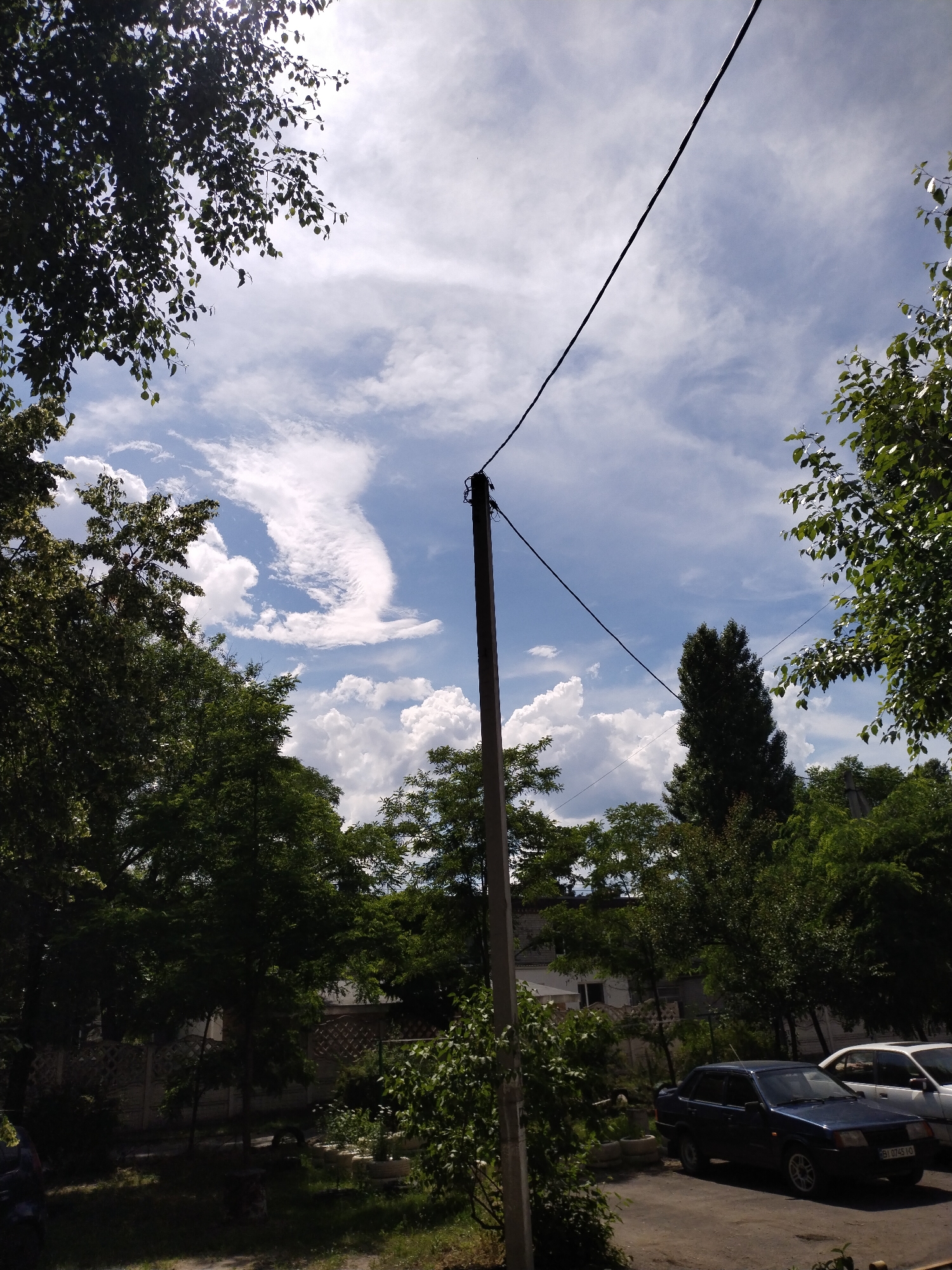 The image depicts an outdoor scene in Horishni Plavni, Ukraine. The viewpoint is directed upwards towards a bright, cloudy sky, with a power line extending diagonally across the frame from the upper left to the lower right. Lush green trees with dense foliage frame the sky on the left and right sides of the image, with sunlight filtering through the leaves on the upper left. In the mid-ground and lower portion of the image, a residential area is visible, characterized by a grey fence with a decorative pattern, some buildings, and a parking area. Two cars are parked: a dark blue sedan in the foreground and a white sedan behind it. The ground appears to be a mixture of dirt and gravel. The overall impression is a daytime scene in a semi-urban or suburban environment.
