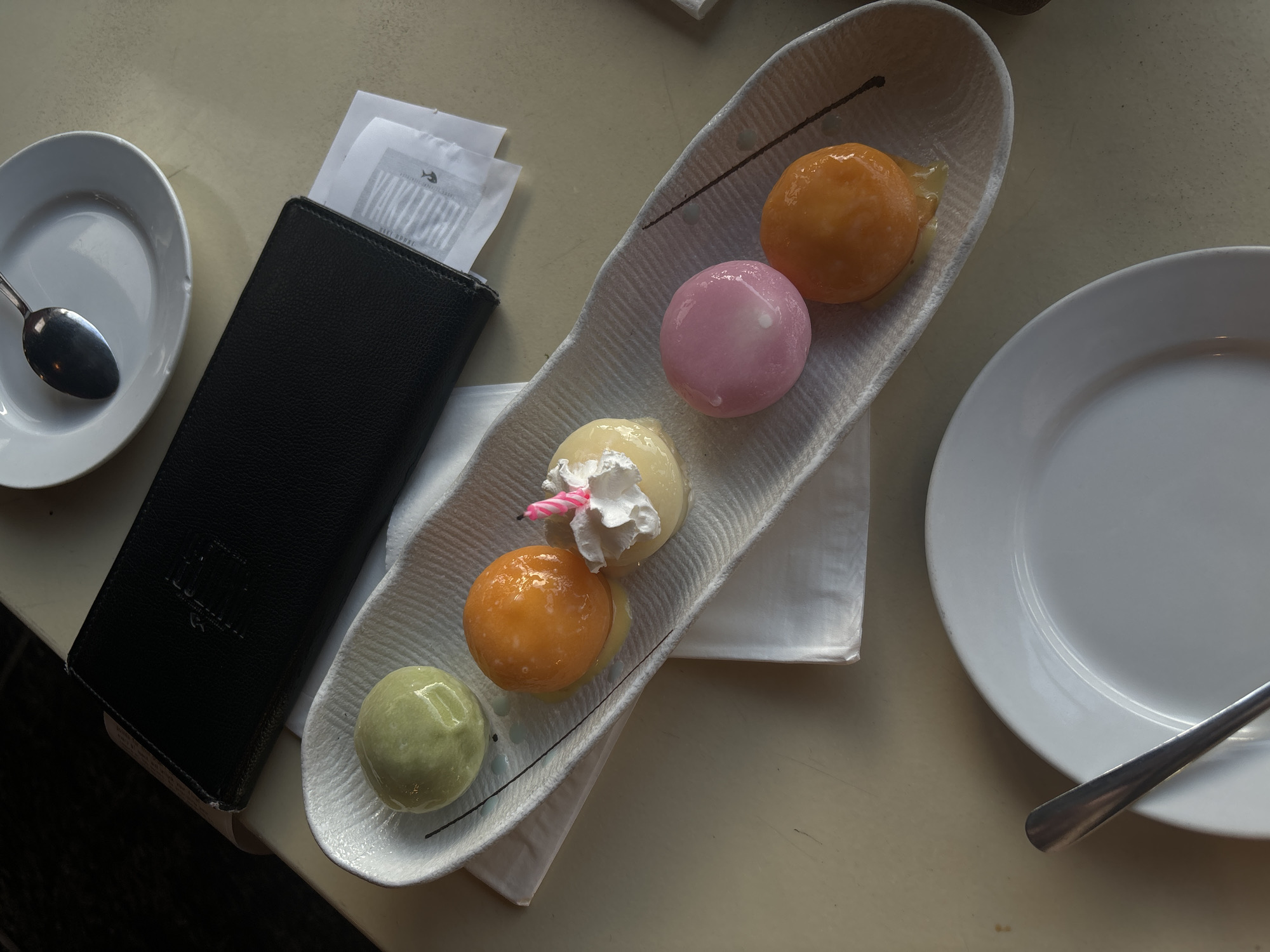 An overhead, close-up shot shows a dessert with five round, pastel-colored mochi on a white, textured serving dish with a black pen. The desserts are arranged in a line with the leftmost being a light green mochi, followed by an orange mochi, then a small pale yellow mochi topped with whipped cream and a pink birthday candle. The next mochi is pink, and the rightmost is orange. All mochi are coated in a shiny glaze, and the orange mochi on the right appears to be melting into a liquid. A black leather menu is placed behind the dessert dish, with a small white paper receipt peeking out from underneath it. To the right of the menu, a white plate with a silver spoon is visible. To the bottom left of the serving dish, a white plate with a silver spoon is also visible. The table is a light beige color and appears to be made of wood. The lighting suggests it is daytime, possibly with natural light coming from a window. The scene is indoors, in a restaurant setting, likely in Boca Raton, Florida, United States of America.