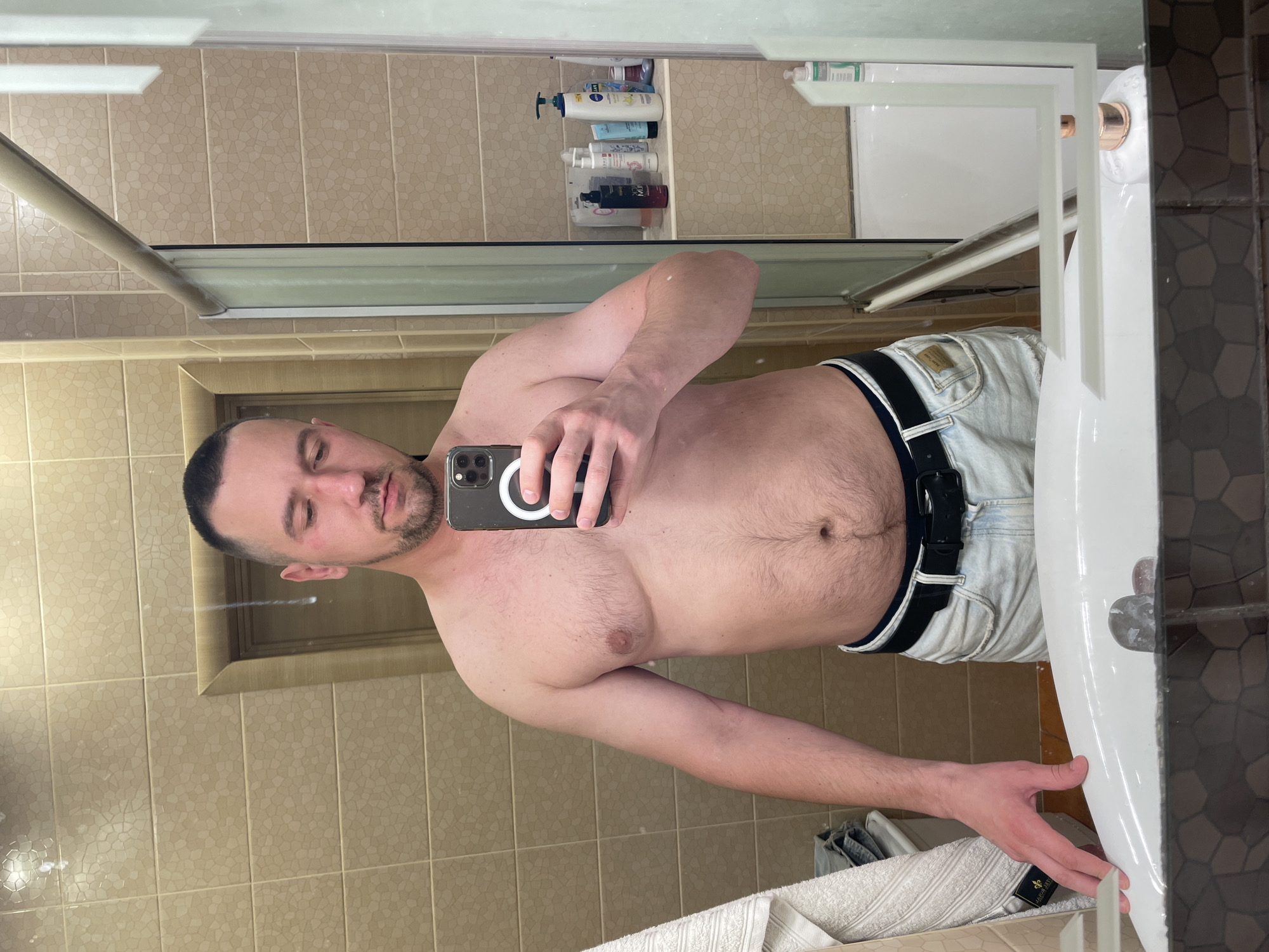 A man is taking a mirror selfie in a bathroom. He is shirtless and wearing light-wash denim shorts with a black belt. The bathroom has beige tiled walls and a white sink. In the background, a shower door is visible, along with shelves containing various toiletries. The man is holding a smartphone in his right hand, angled to capture the reflection. The setting is a domestic bathroom in Kyiv, Ukraine.