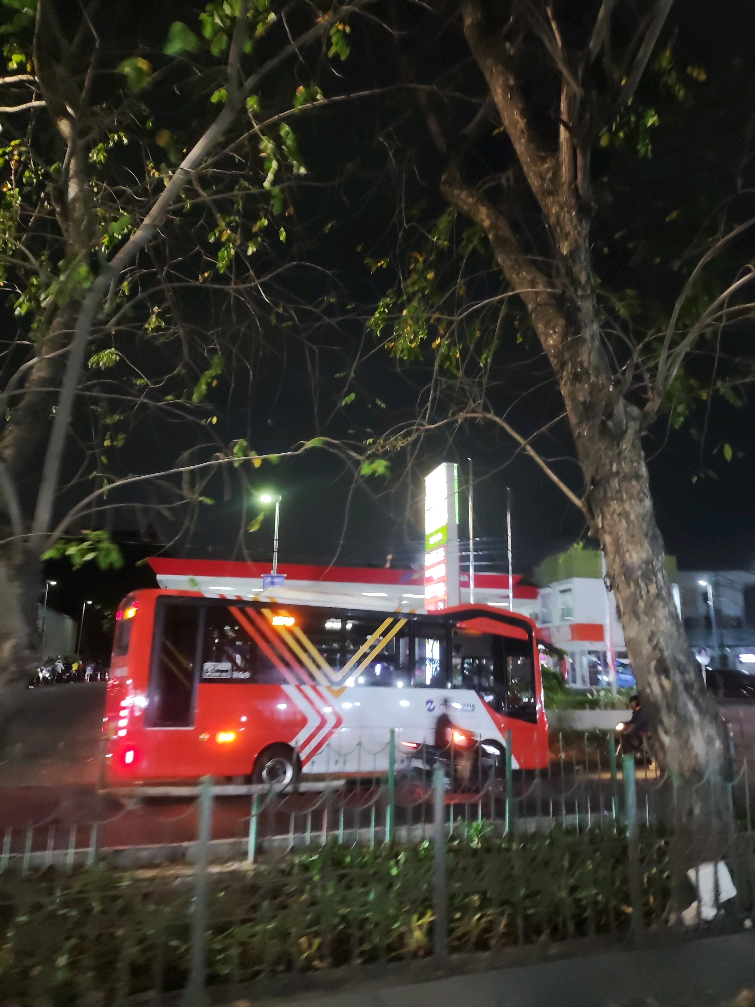 This image depicts a street scene at night in East Jakarta, Indonesia, likely near a gas station. A red and white public bus with illuminated taillights and side indicators is the central subject, moving on a road. The bus has a distinctive diagonal stripe pattern in yellow and white. In the background, a brightly lit gas station with multiple pumps is visible, along with a white building with red accents. Trees with sparse leaves flank the scene, with branches extending over the road and obscuring parts of the sky. The ground appears to be asphalt or a similar road surface, with a metal fence and some greenery in the foreground. A few individuals on motorcycles are present on the left side of the road, and a person on a motorcycle is seen to the right of the bus. Streetlights are also illuminating the area. The overall impression is a typical urban night scene with a focus on public transportation and commercial infrastructure.
