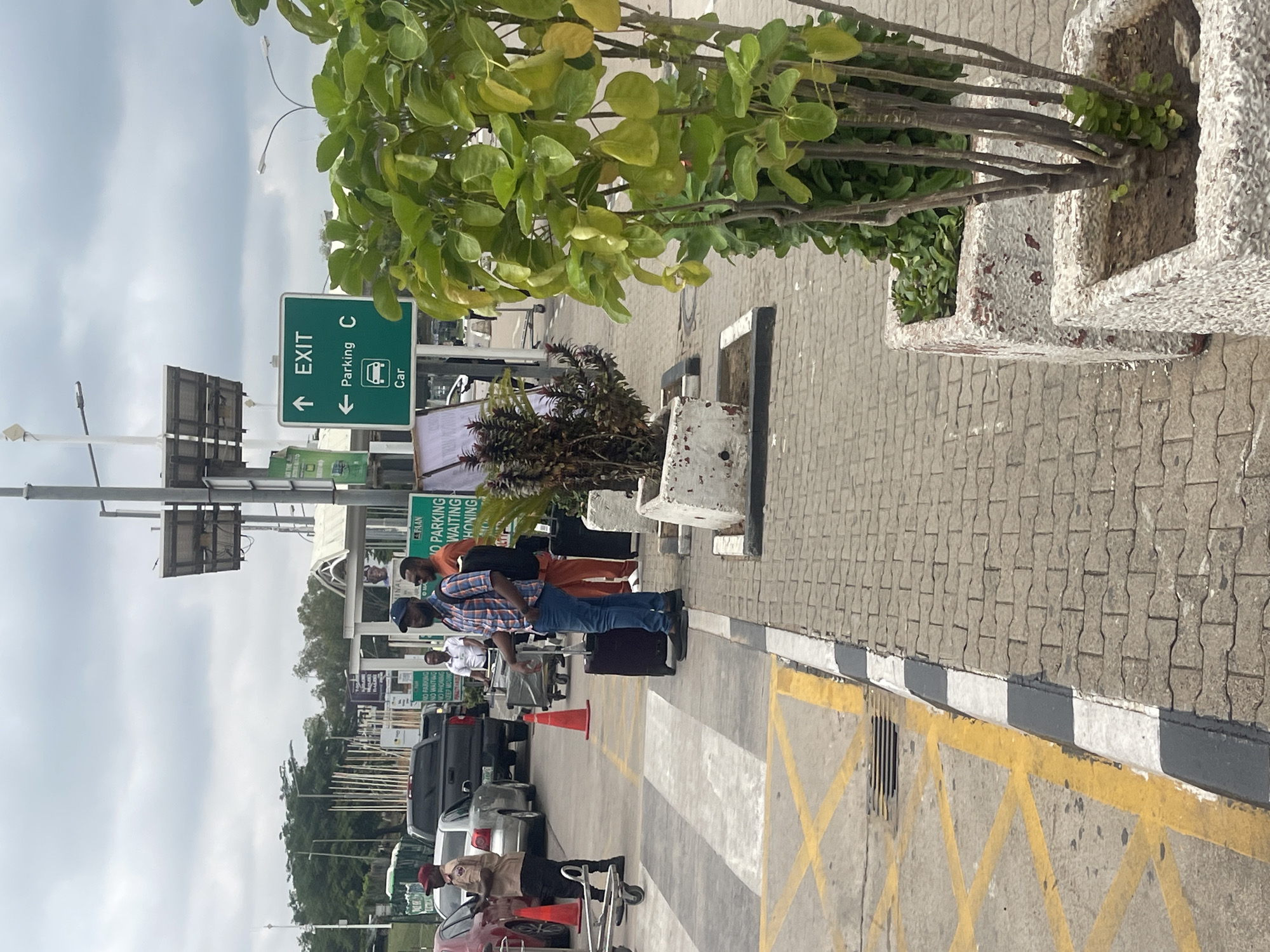 The image, rotated 90 degrees counter-clockwise, depicts an outdoor scene at what appears to be an airport or a transport hub, likely in Gwaska, Nigeria. Several people are visible, some standing with luggage, suggesting they are travelers waiting or moving through the area. Cars are parked or waiting in the background.
The setting includes a paved walkway with a prominent black and white striped curb separating it from a vehicle area marked with yellow crosshatch patterns. An orange traffic cone is also visible in the vehicle area. Lush green plants in concrete planters line the pedestrian zone. The sky is overcast, indicating a cloudy daytime.
Visible signage provides specific location cues: A green sign clearly displays "↑ EXIT" and "← Parking C" with a "Car" label and an icon of a bus or vehicle. Another sign, partially obscured, includes the acronym "FAAN" (Federal Airports Authority of Nigeria) and the phrases "NO PARKING," "WAITING," and "HONING" (likely "HONKING" misspelled or abbreviated).