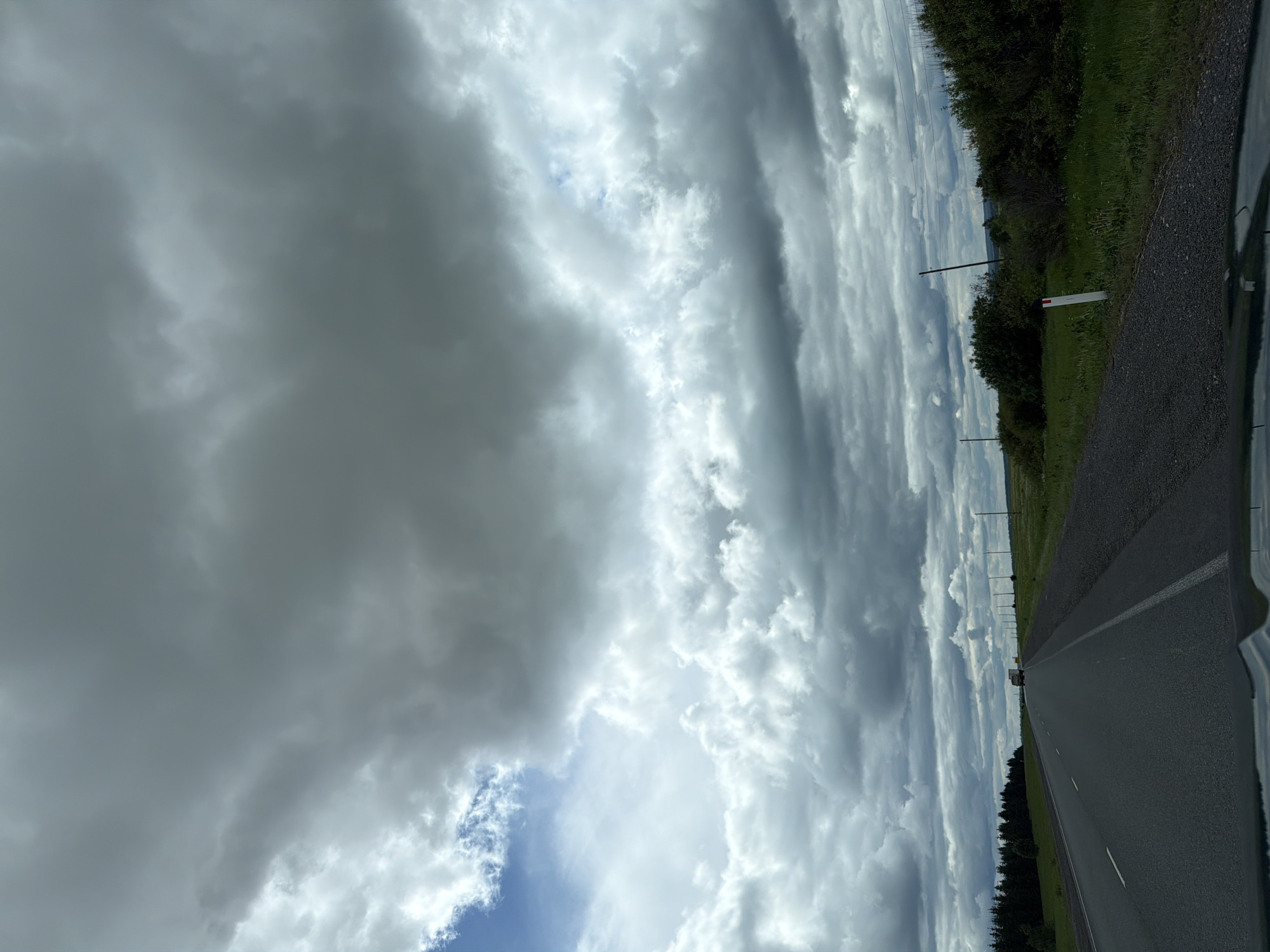 The image depicts a road in Горняк, Russia, under a sky filled with dramatic, layered clouds. The road is asphalt and appears to be a two-lane highway. The shoulder of the road is lined with green grass and vegetation. A few utility poles are visible along the roadside. The sky is dominated by various cloud formations, with some patches of blue sky peeking through.