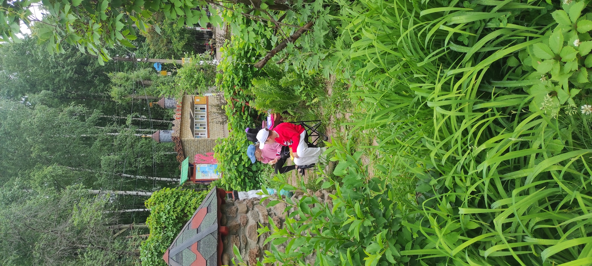 Two people, appearing to be children, are sitting on folding chairs in a garden. The person on the left is wearing a pink sweater and a blue baseball cap. They are holding something in their hands and appear to be eating. The person on the right is wearing a red sweatshirt and a white baseball cap. They are also holding something in their hands. In the background, there are small, decorative buildings that resemble castles, and a larger stone structure with a red and green roof on the left. The area is filled with lush green vegetation, including tall grasses and leafy plants. The scene suggests a park or recreational area, possibly a children's play area or a themed garden. The location is identified as the city of Chaikovsky, Russia.