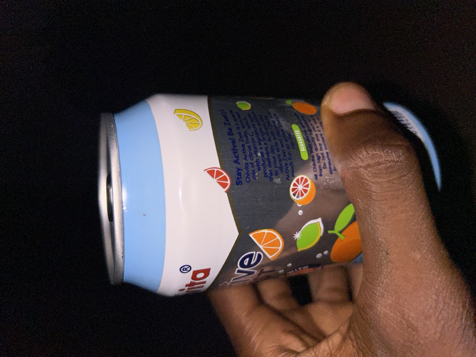 In Egbe, Nigeria, a person's hand with dark skin grips a Chivita Active Zest beverage can against a uniformly dark background. The aluminum can features a light blue top section, transitioning to white, and a dark grey body. Visible text on the can's label includes "Chivita Active" with a registered trademark symbol, the slogan "Stay Active! Be Zesty!", and descriptive text stating, "Chivita Active Zest is a special mix of citrus fruits, enriched with Vitamin C, which produces a signature taste that delivers a bright active to your day." Illustrations of various citrus fruit slices, including oranges, lemons, limes, and grapefruits, are dispersed across the dark grey label. A "GUARANTEE" section is also visible near the bottom of the described portion of the can. The can's pop-top is intact, showing the pull-tab mechanism.