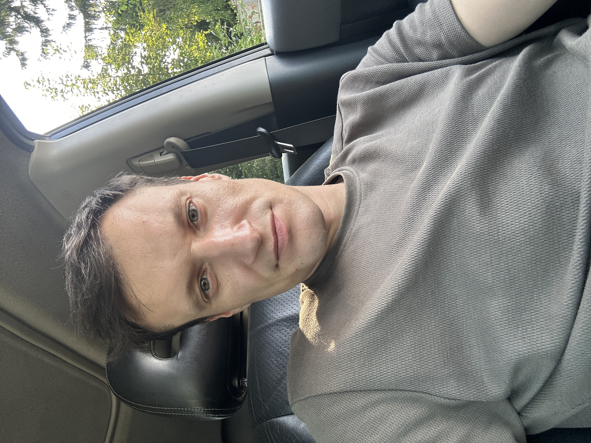 A man with short, dark hair and light eyes is shown in a selfie taken from inside a car. He is wearing a short-sleeved, grey t-shirt. The interior of the car is visible, including a black leather seat and a portion of the car's roof. Outside the car window, trees and foliage are visible under a bright sky. The image is a head-and-shoulders shot, with the man looking directly at the camera. The sun is casting a shadow on his left shoulder and shirt. The specific location in Shatava, Ukraine is not discernible from the visual information provided in the image.