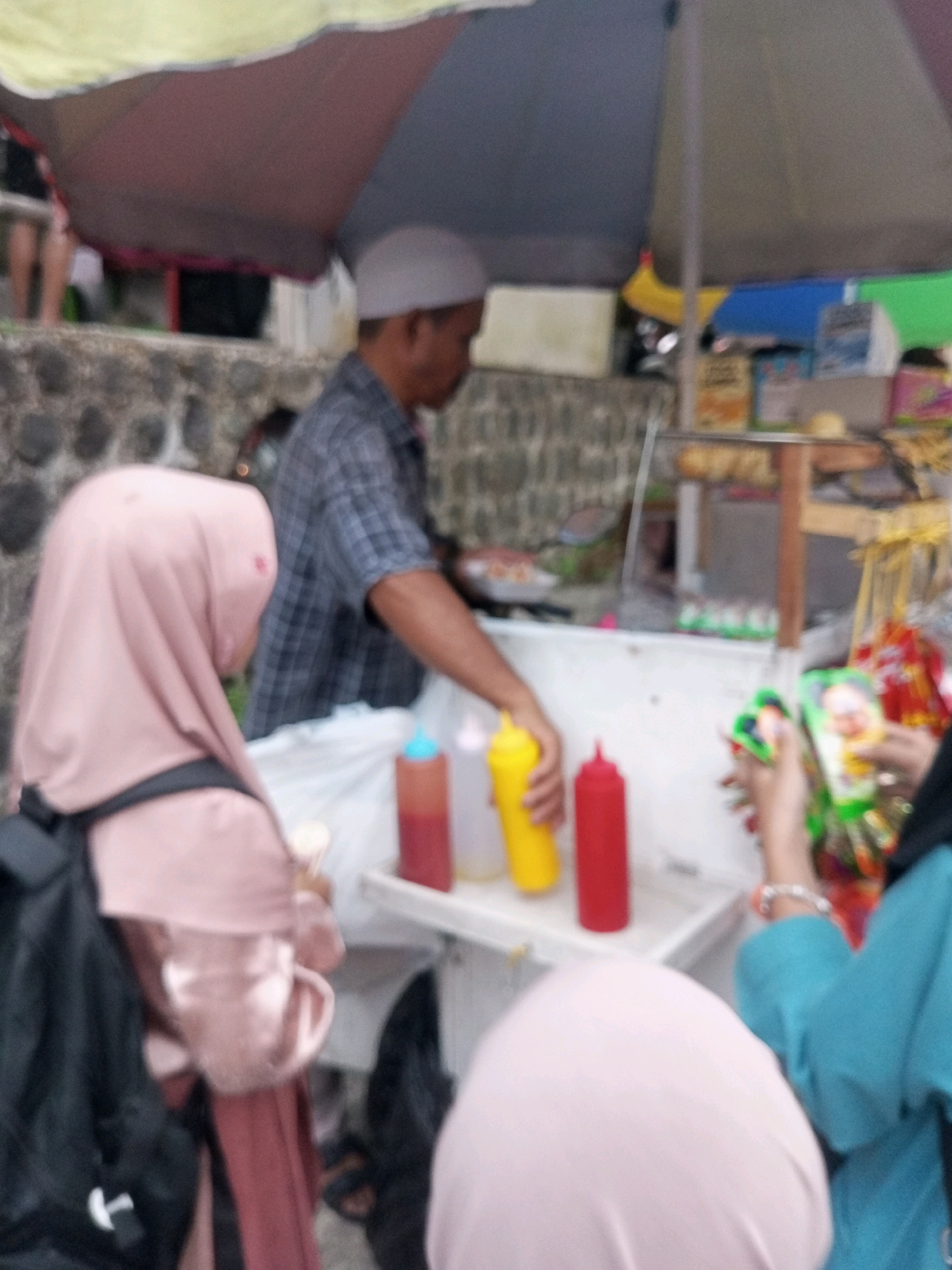 The image captures a street vendor scene in Krasak, Indonesia. In the foreground, a person wearing a pink hijab is partially visible, facing away from the camera. To their right, a vendor is serving a customer from a stall. The vendor, wearing a checkered shirt and a white cap, is handling bottles of condiments. The stall is covered by a large umbrella and displays various food items and packaged snacks. In the background, a stone wall and other structures suggest an urban or semi-urban environment. The overall atmosphere appears to be that of a busy local marketplace.