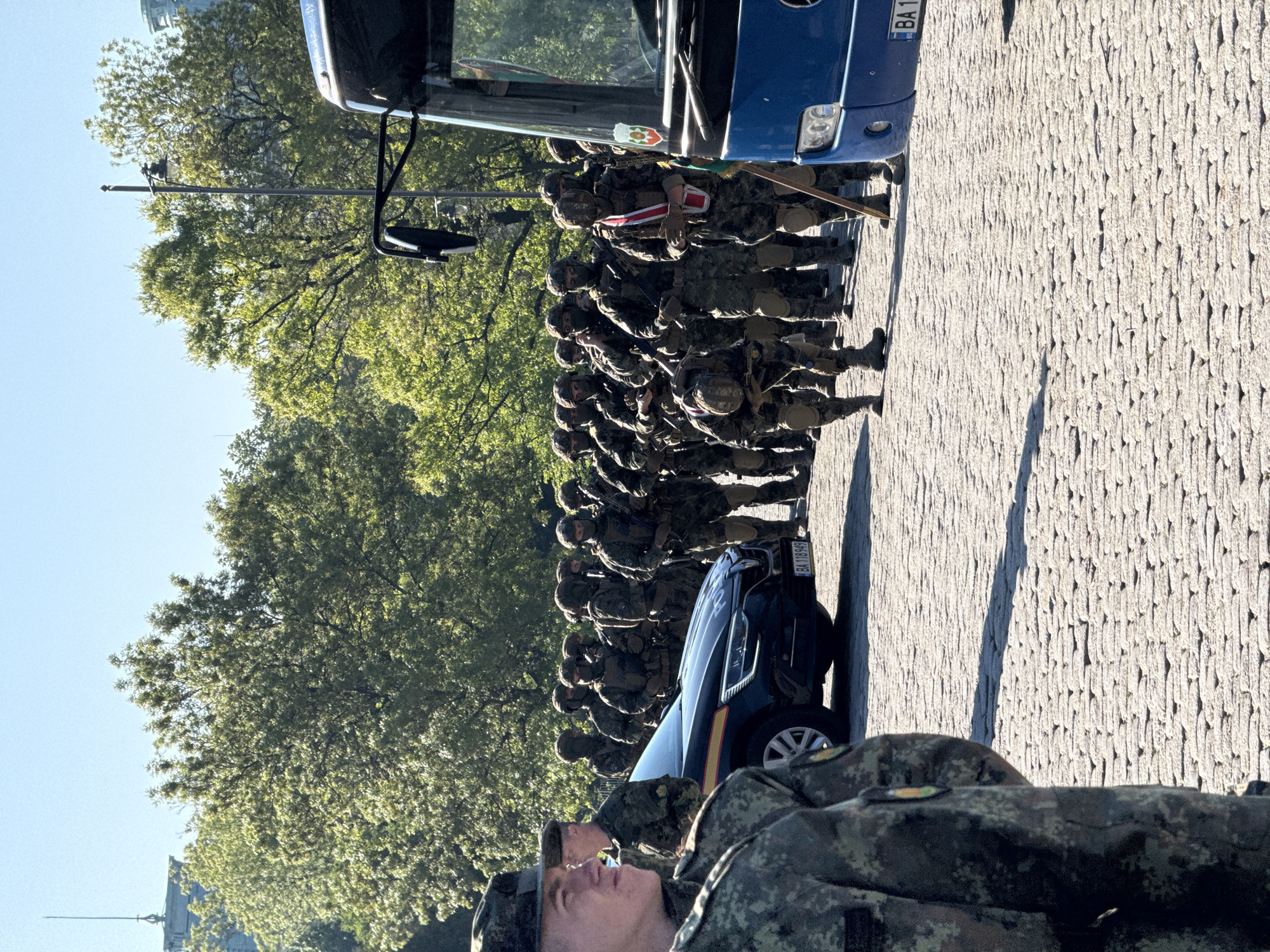 The image depicts a group of soldiers standing at attention on a cobblestone street in Sofia, Bulgaria. They are lined up near a blue bus and a police car, both of which have license plates starting with "BA". The soldiers are in camouflage uniforms, and some are holding rifles. The setting appears to be an urban area with trees and buildings in the background.