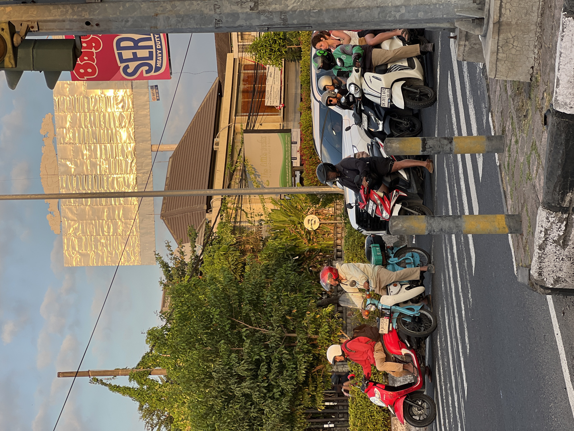In Denpasar Barat, Indonesia, a group of motorcyclists waits at a pedestrian crossing during what appears to be the golden hour. The sky is partly cloudy, with soft sunlight casting a warm glow. In the foreground, traffic lights and a concrete barrier are visible. Several motorcycles are stopped, with riders and passengers wearing helmets. A red scooter, a light blue scooter, and several black and white scooters are present. The riders are dressed casually, in t-shirts and shorts, with some wearing long pants. The background features lush green trees, buildings, and a large billboard advertising "SER HEAVY DUTY." The overall scene depicts a common urban streetscape in Indonesia.